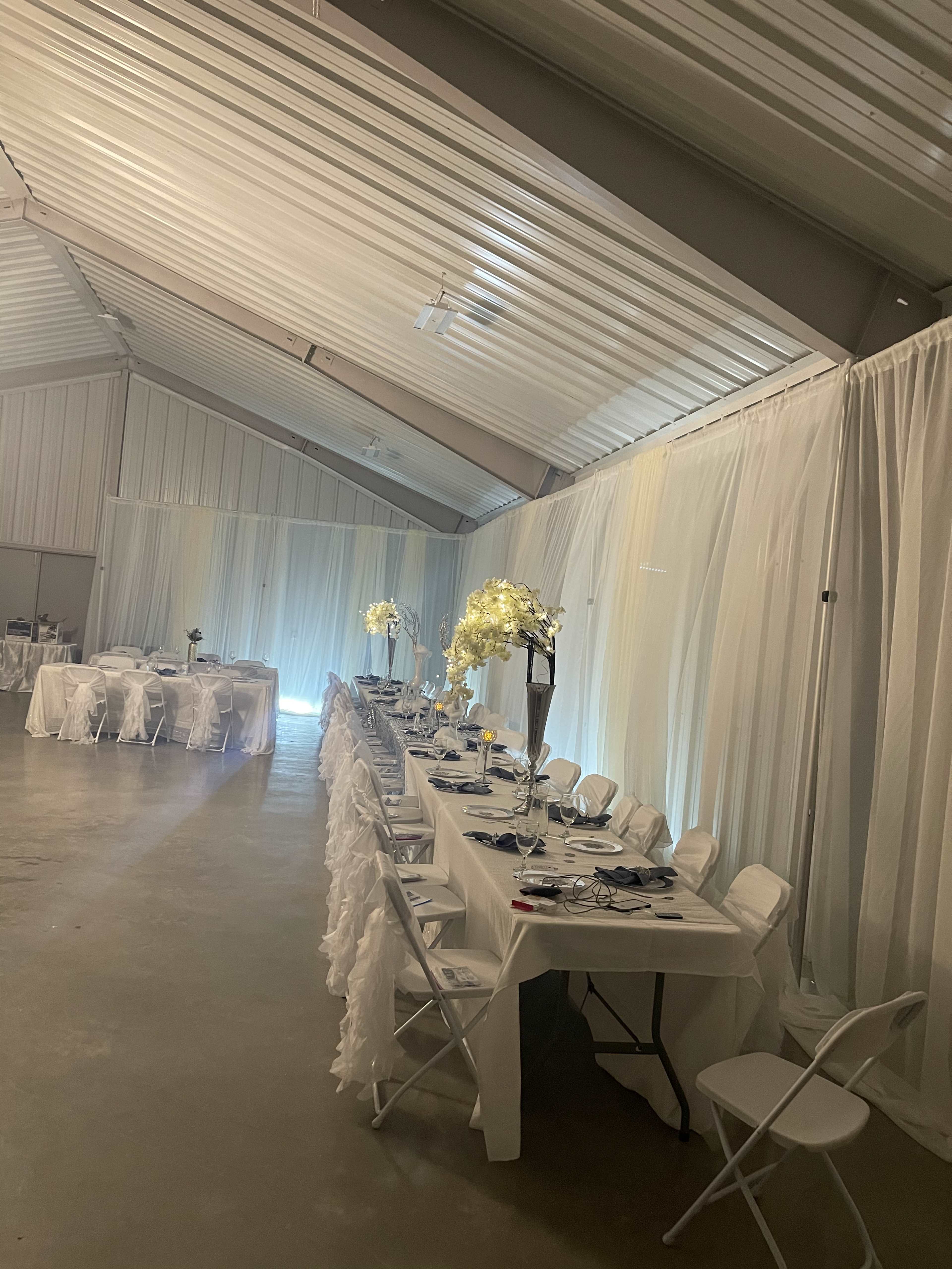 A long banquet table is set up in a warehouse-style space, adorned with white linens and floral centerpieces, while chairs are arranged around it.