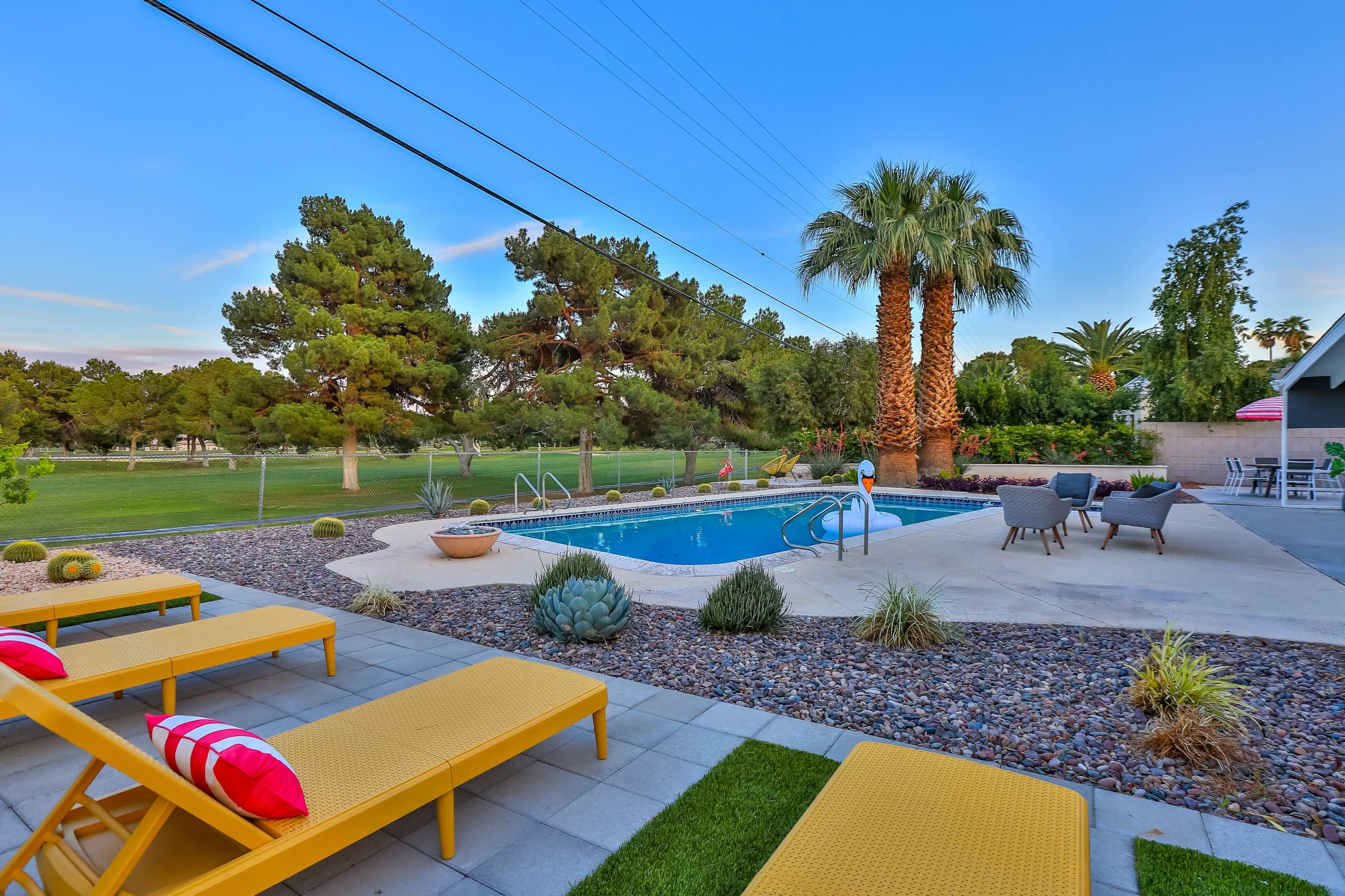A backyard setting featuring a swimming pool surrounded by lounge chairs, palm trees, and landscaped greenery.