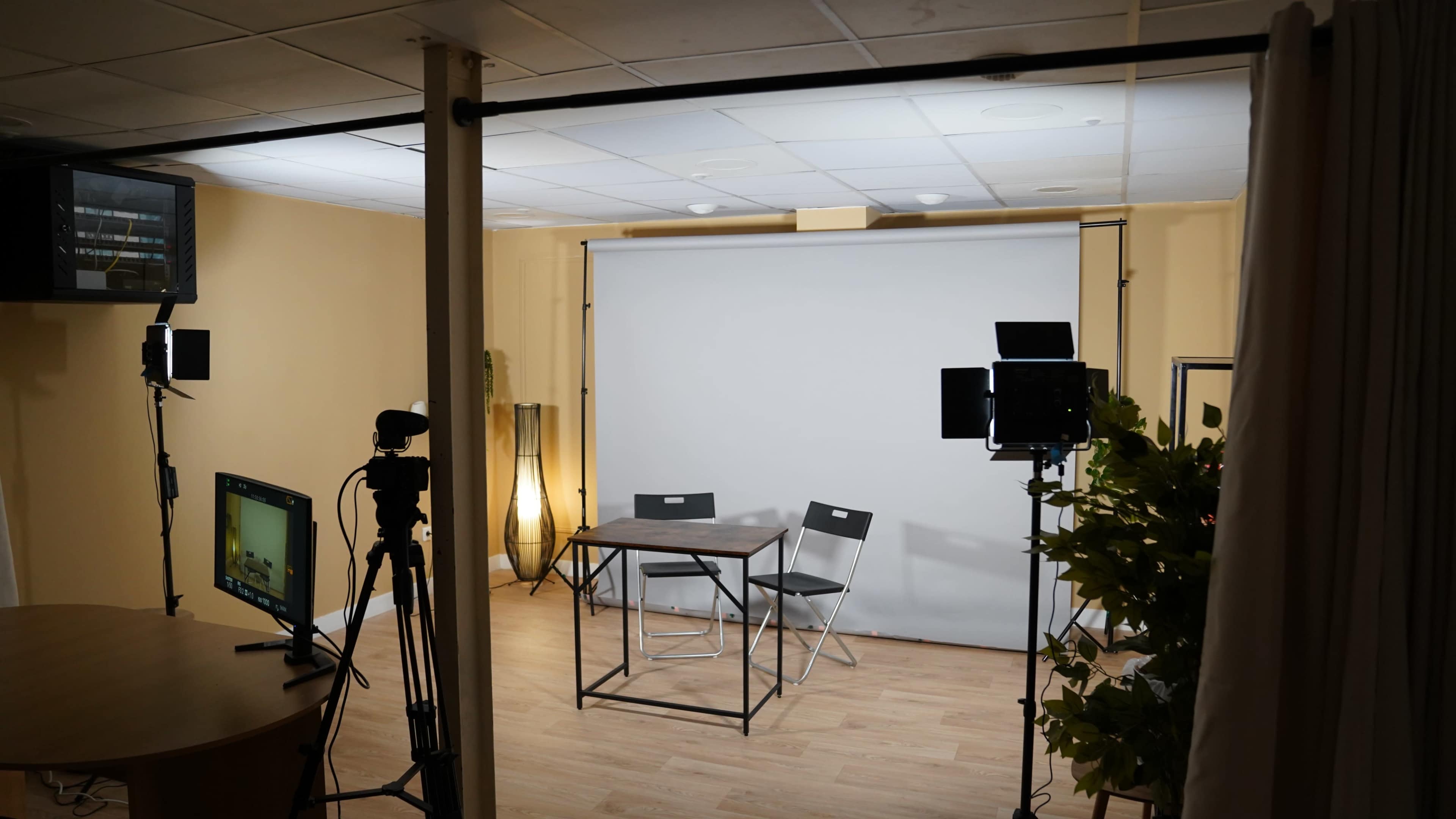 The image shows a minimalistic indoor studio setup with a table, two chairs, and lighting equipment positioned around a blank backdrop.