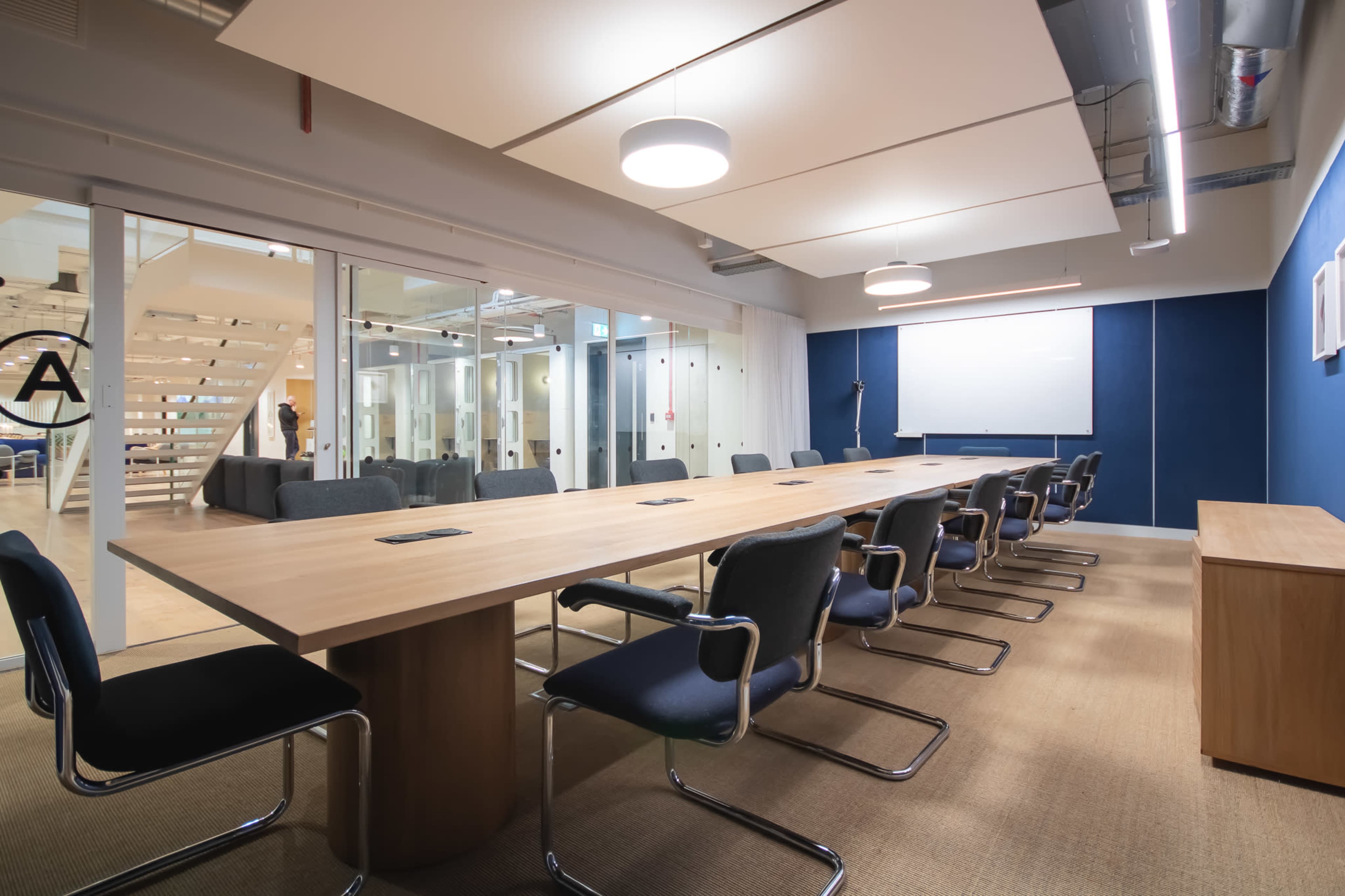 A spacious conference room features a long wooden table surrounded by chairs, with a projector screen on one wall and glass partitions allowing views of adjacent areas.
