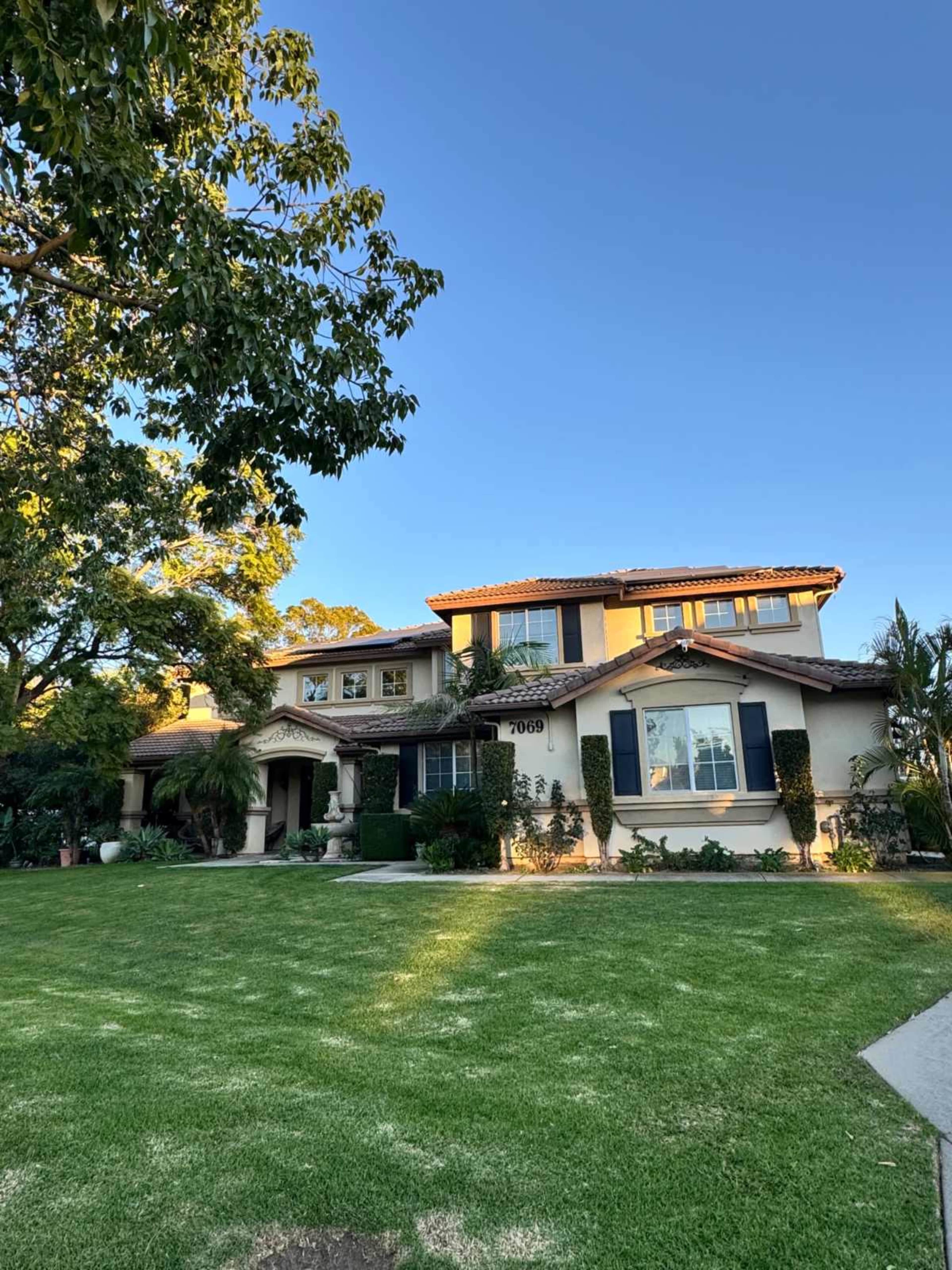 A two-story house with a tile roof and manicured lawn is surrounded by trees and shrubs.