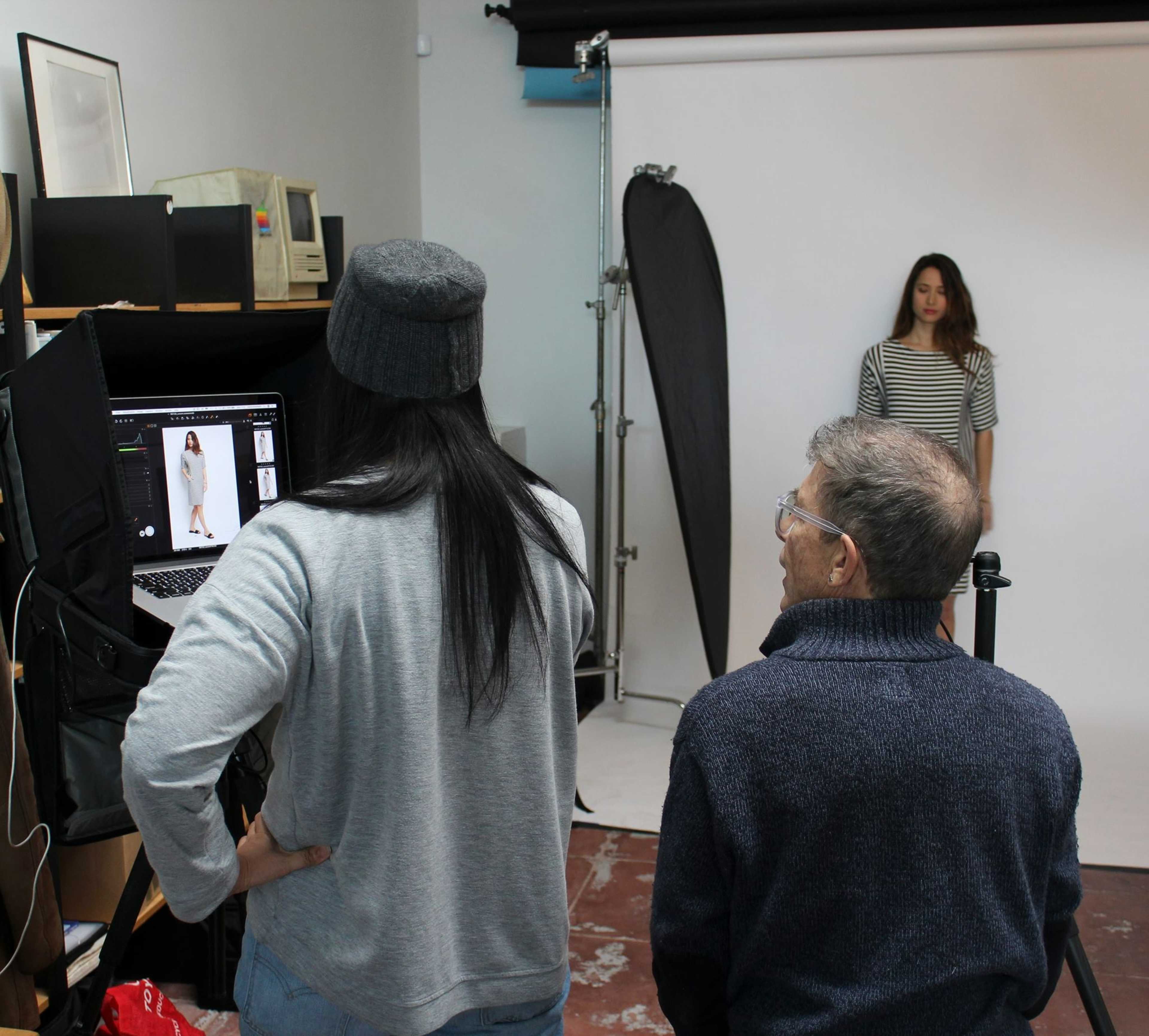 A photographer and a client observe a model posing in front of a backdrop while reviewing images on a laptop in a studio setting.