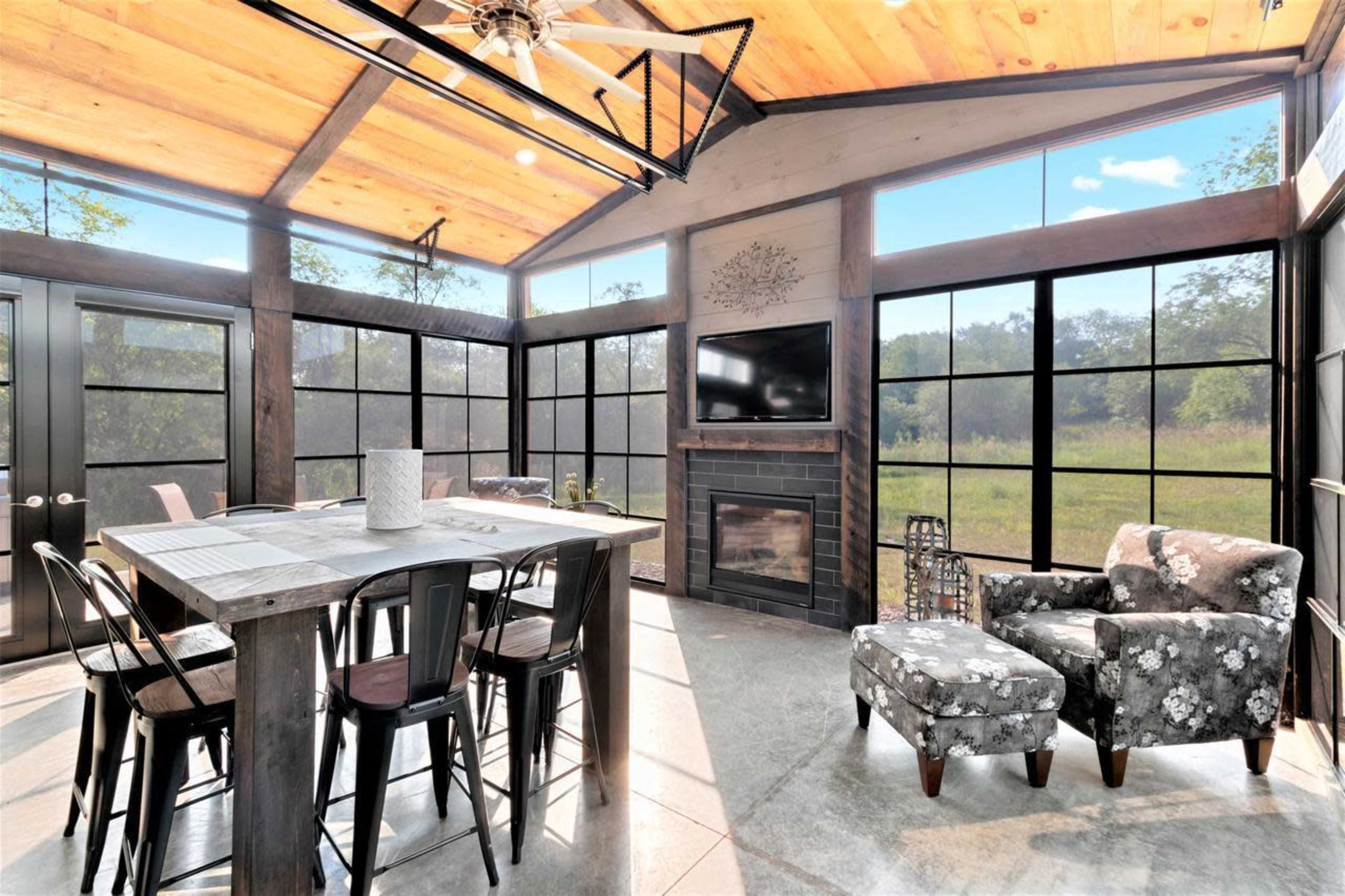 A modern, airy living space features a large dining table, floral-patterned armchair, and a fireplace, with windows offering views of the outdoors.