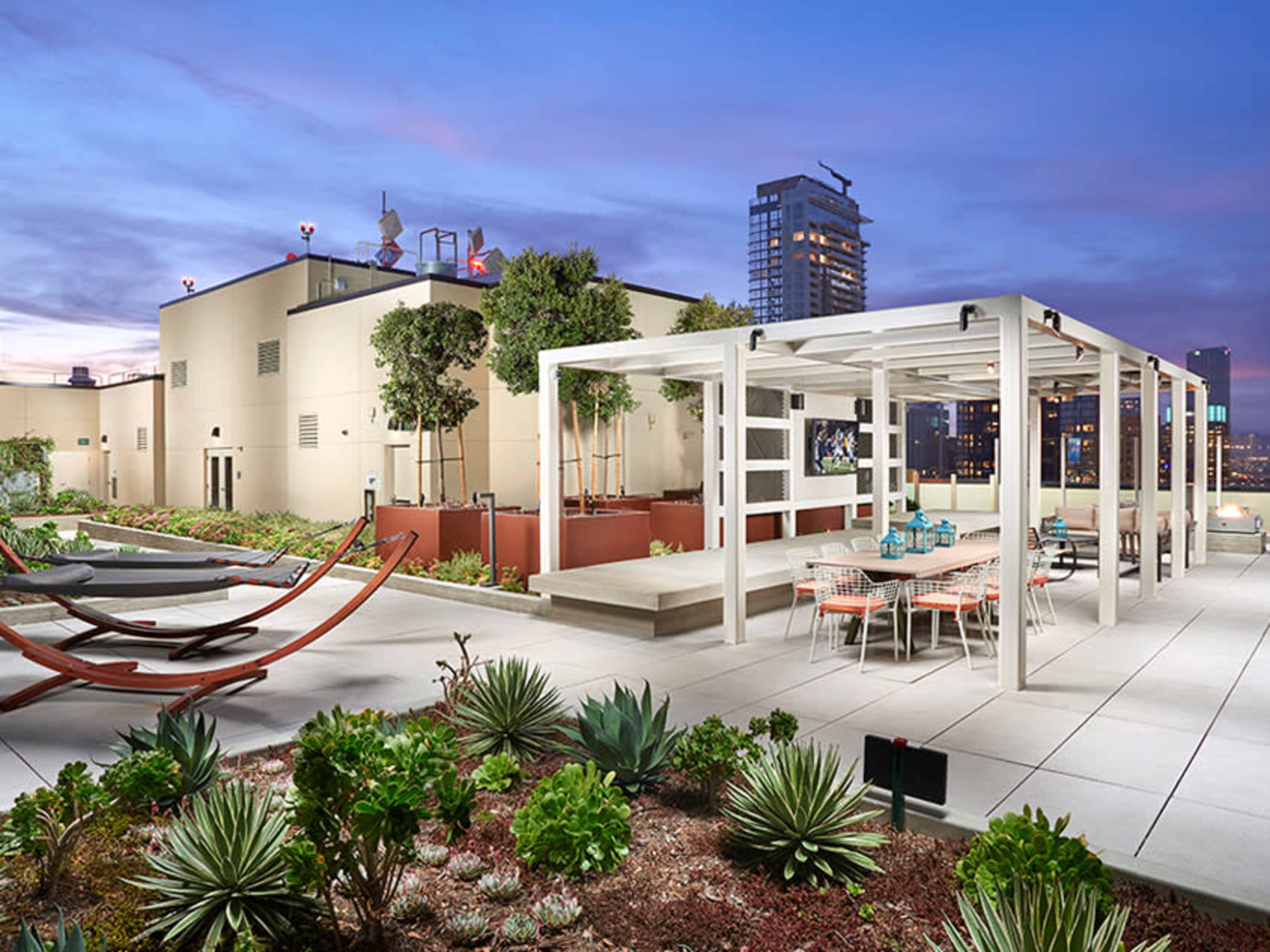 A rooftop terrace featuring a pergola, seating area with tables and chairs, and landscaped plants under a twilight sky.
