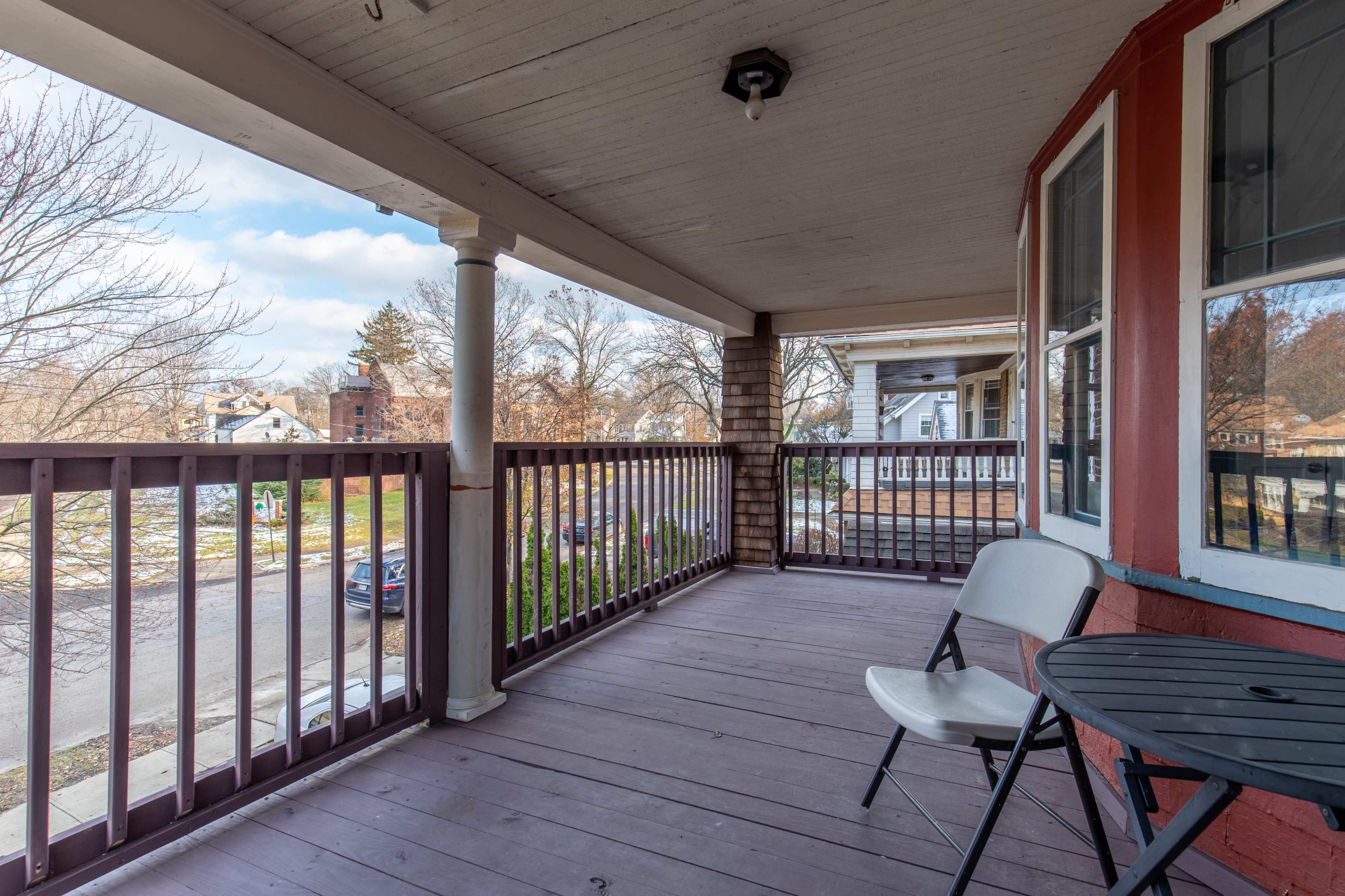 A porch with a small round table and a folding chair, overlooking a street with trees and houses in the background.
