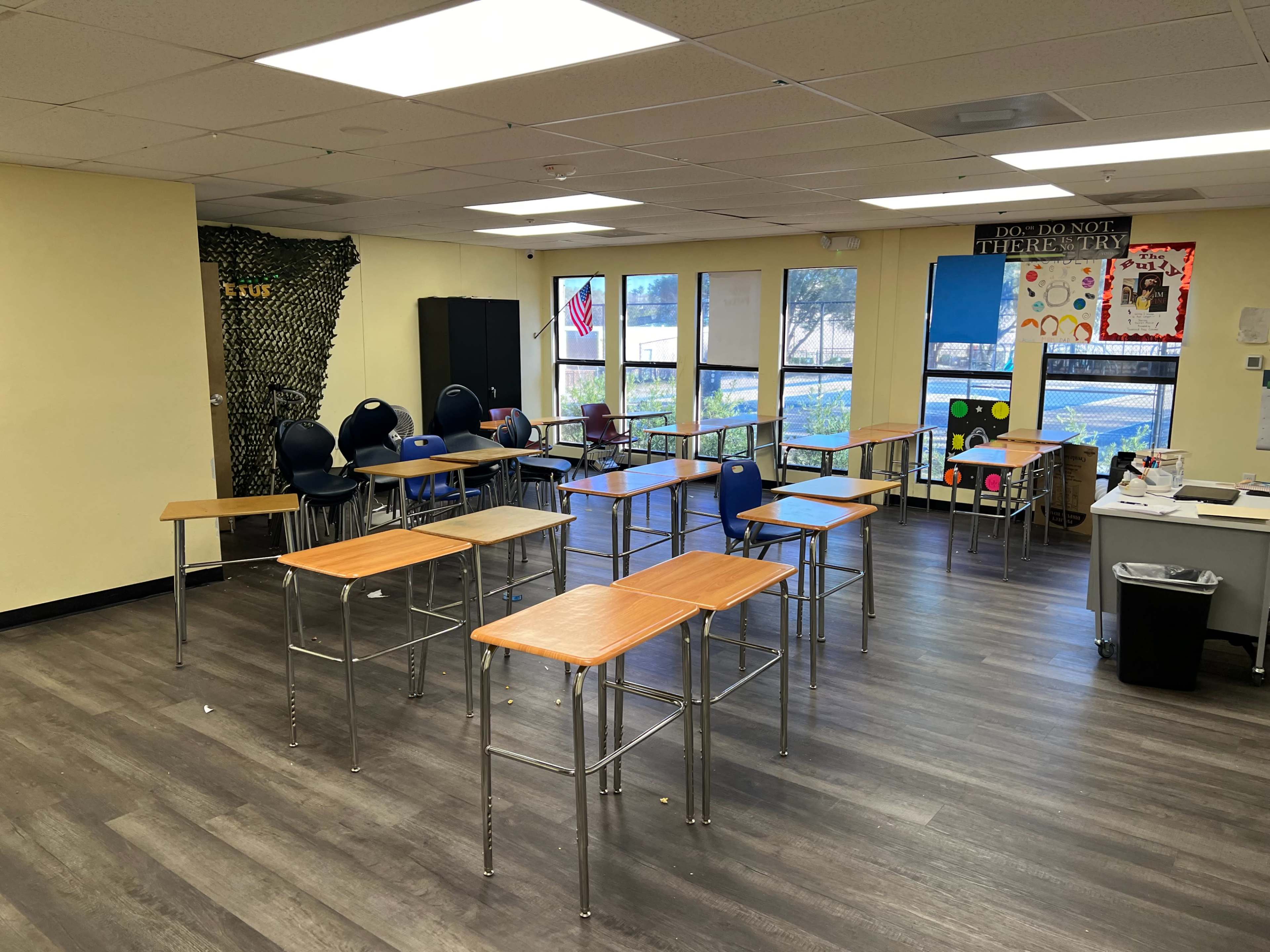 A classroom with several empty desks arranged in a semi-circular layout and large windows allowing natural light to enter.