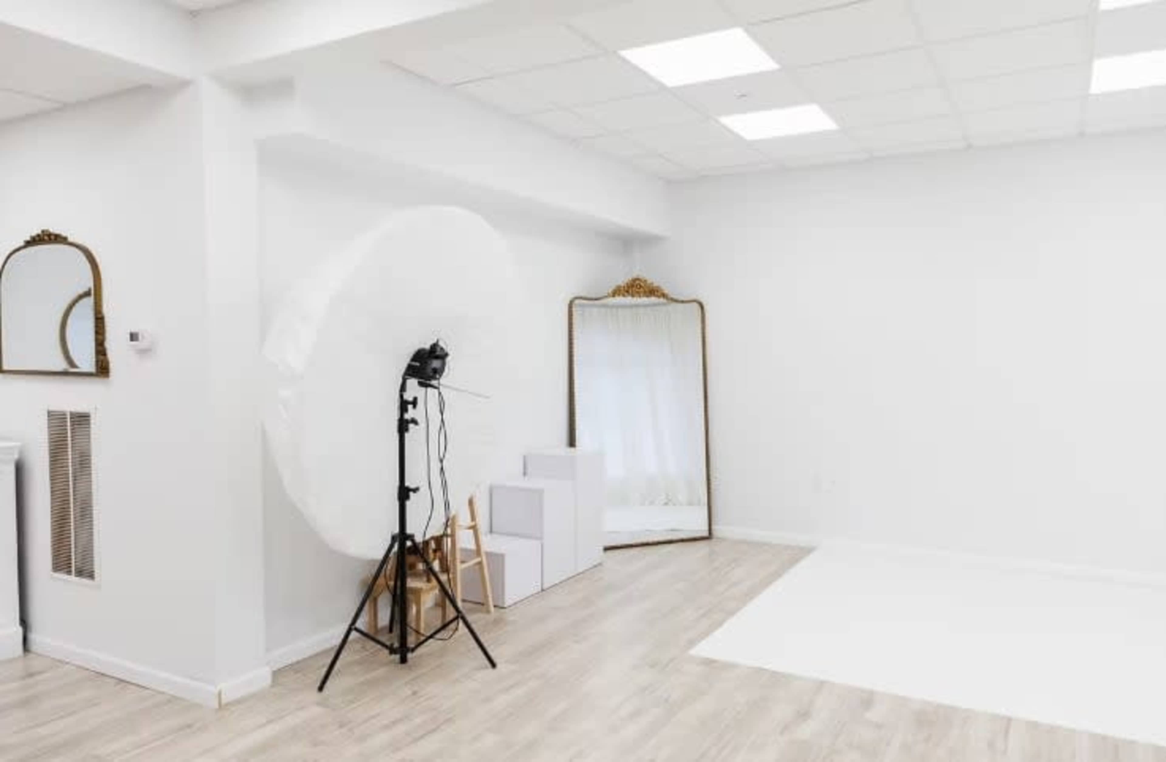 The image shows a minimalist photography studio featuring a large white backdrop, softbox light setup, and an ornate mirror against a clean, white wall.