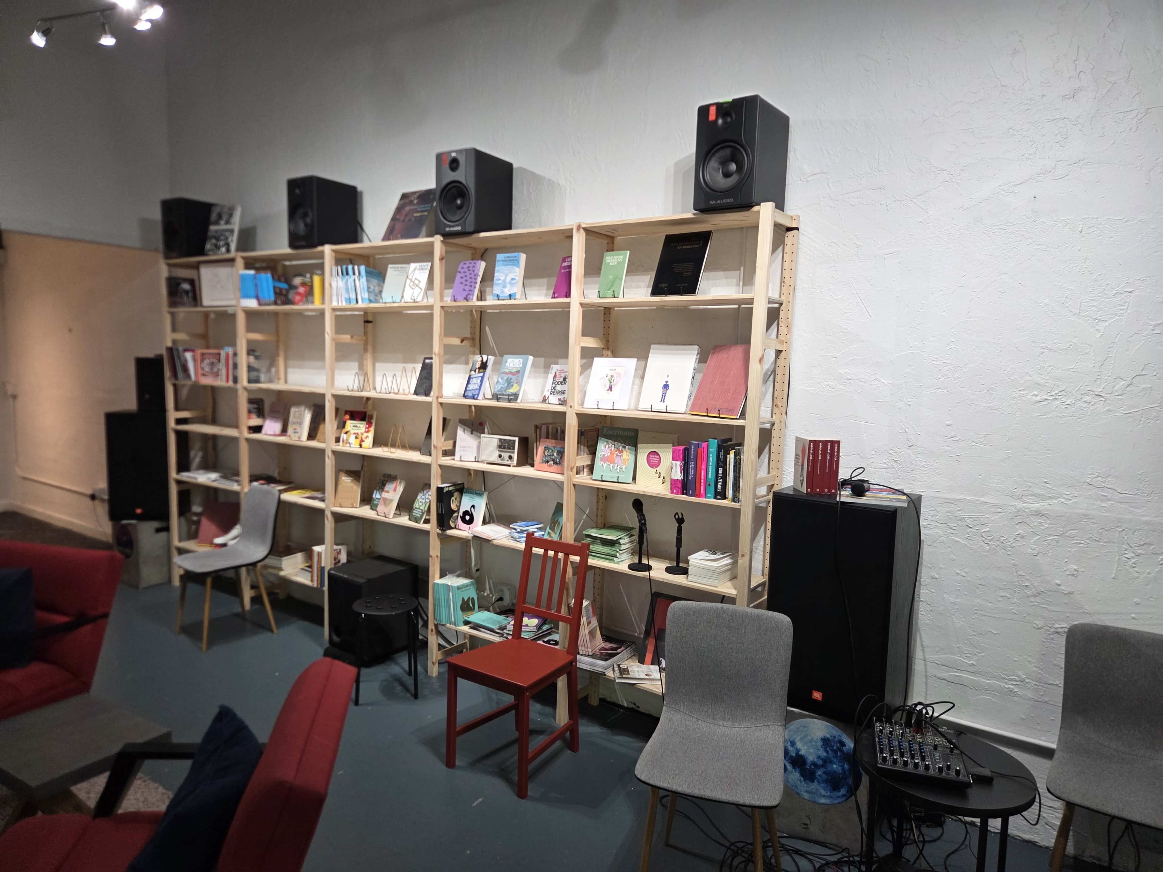 A wooden bookshelf filled with books, magazines, and various items is displayed against a textured wall, with audio equipment and seating in front.