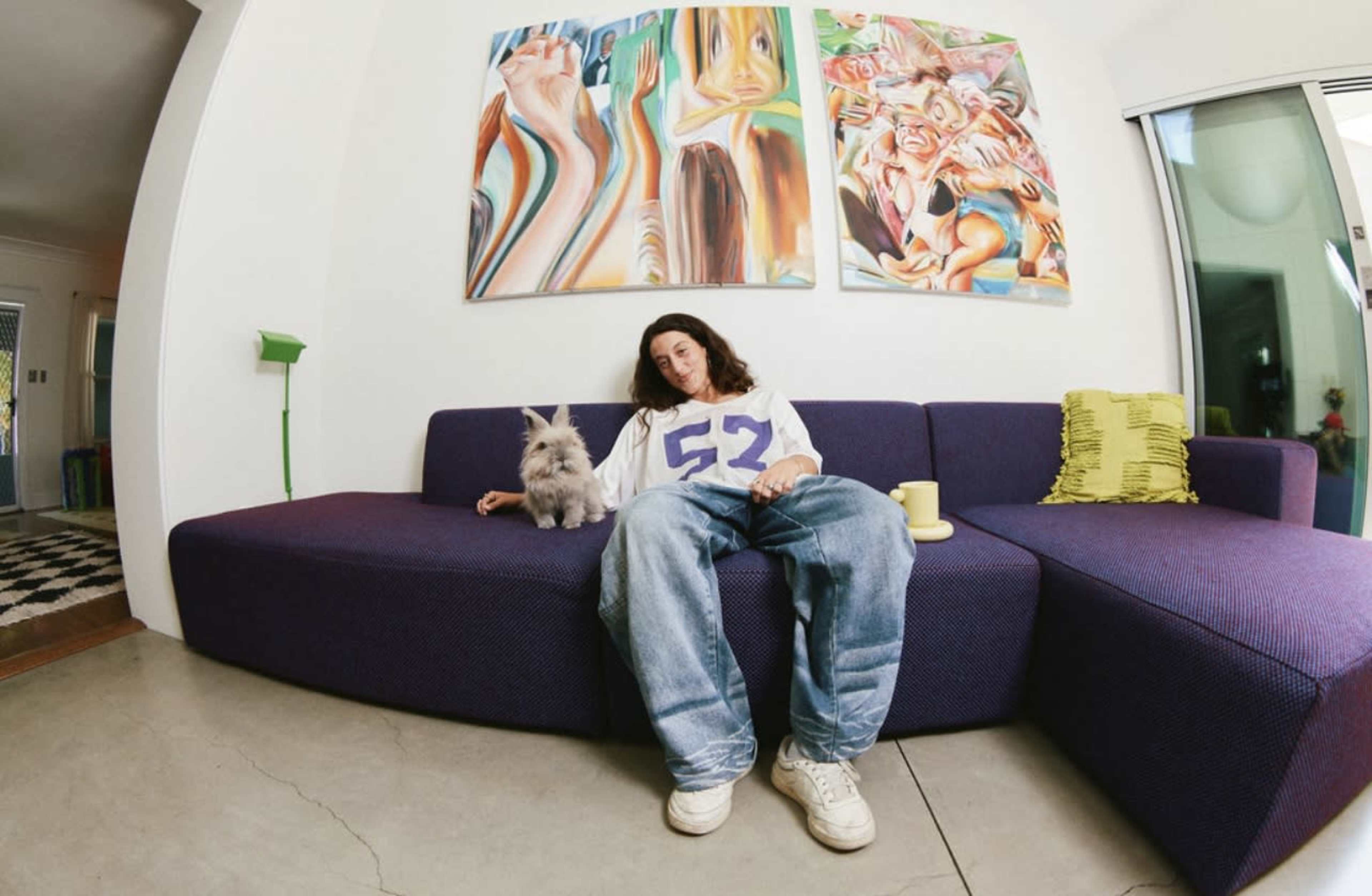 A person sits on a purple couch with a small dog beside them in a room featuring colorful artwork on the walls.