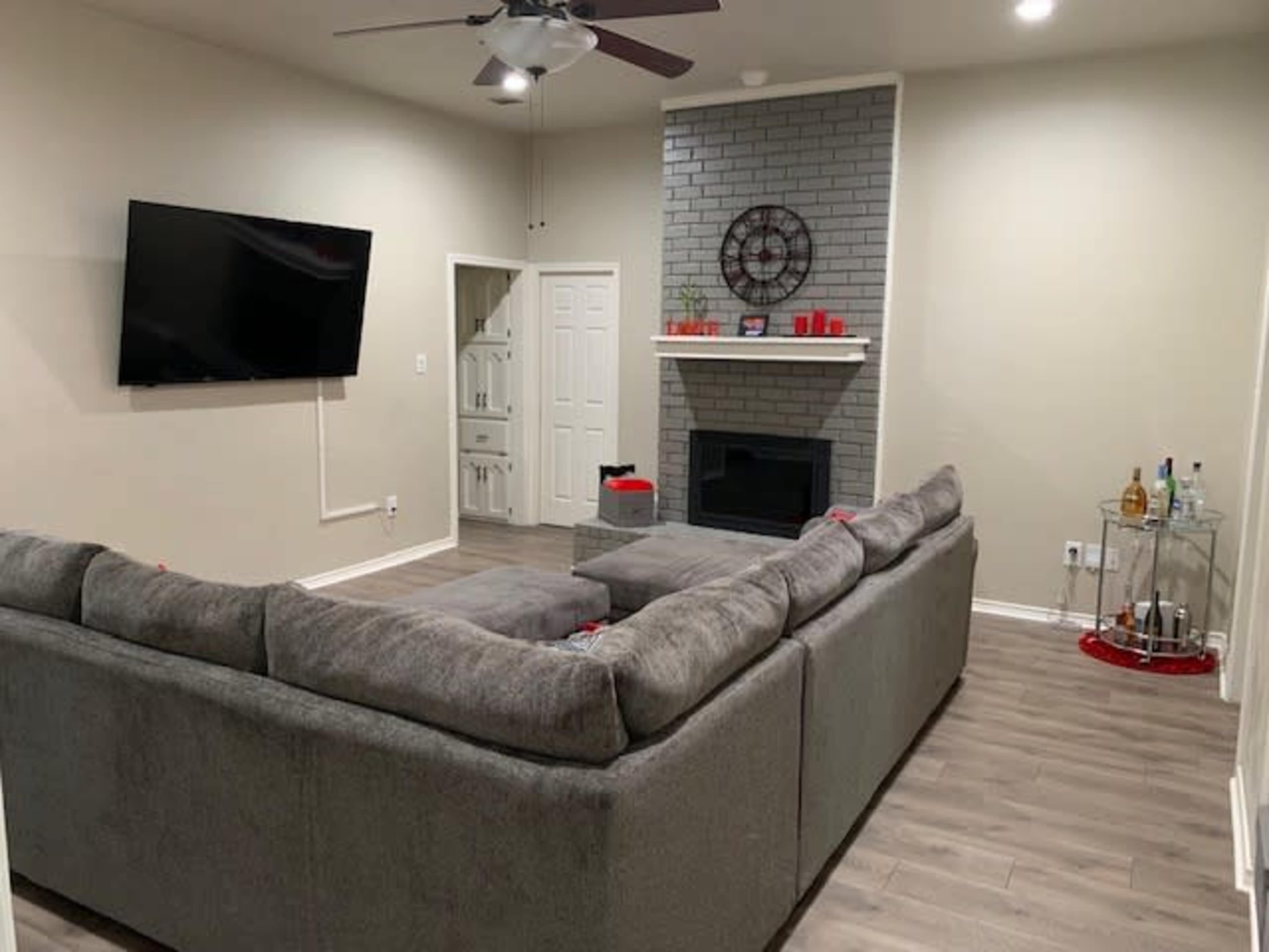A living room with a large gray sectional sofa, a wall-mounted television, a fireplace with a decorative element above it, and a small bar cart in the corner.