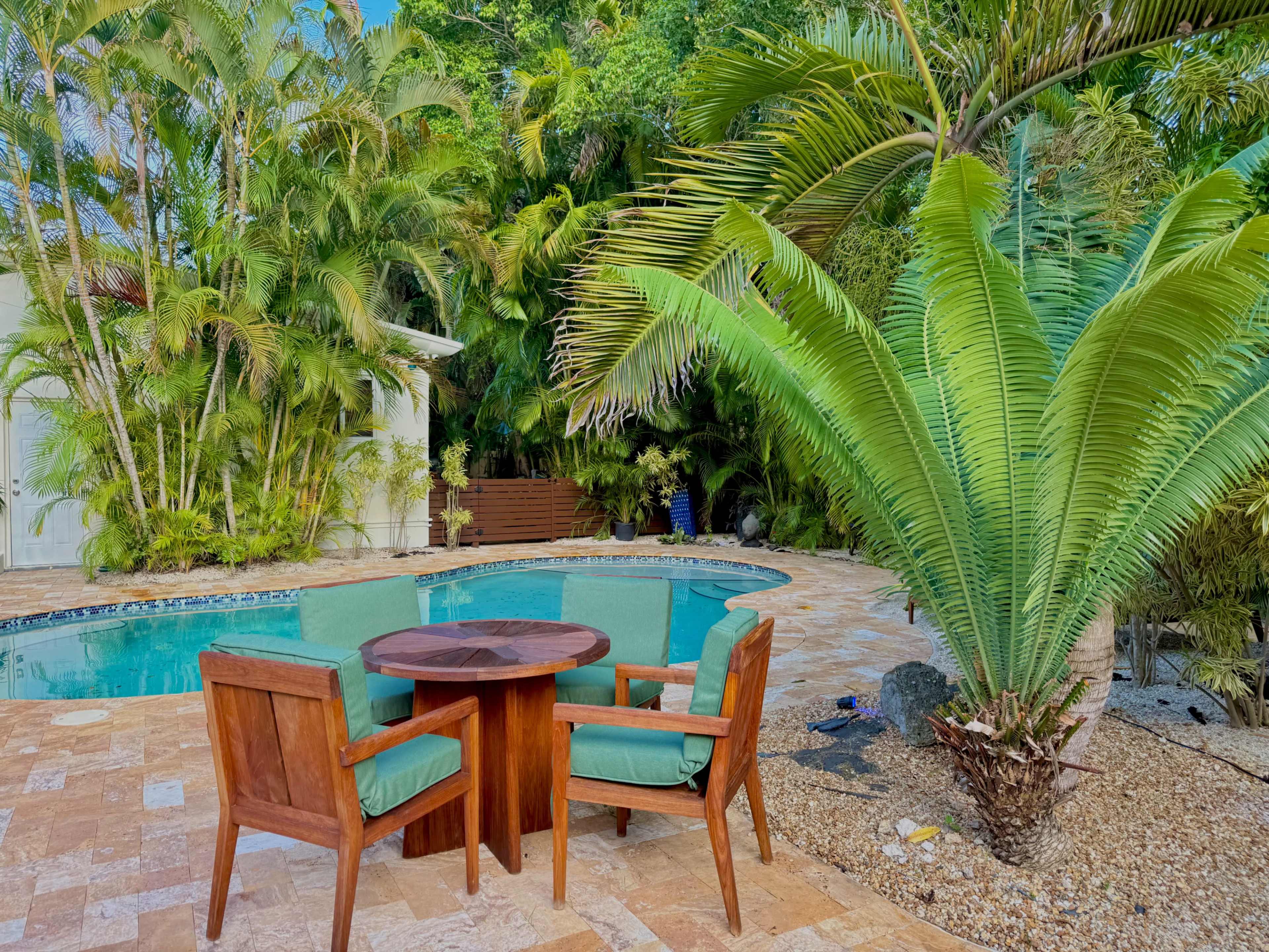 A round wooden table surrounded by four green chairs is positioned beside a swimming pool, framed by lush tropical plants.