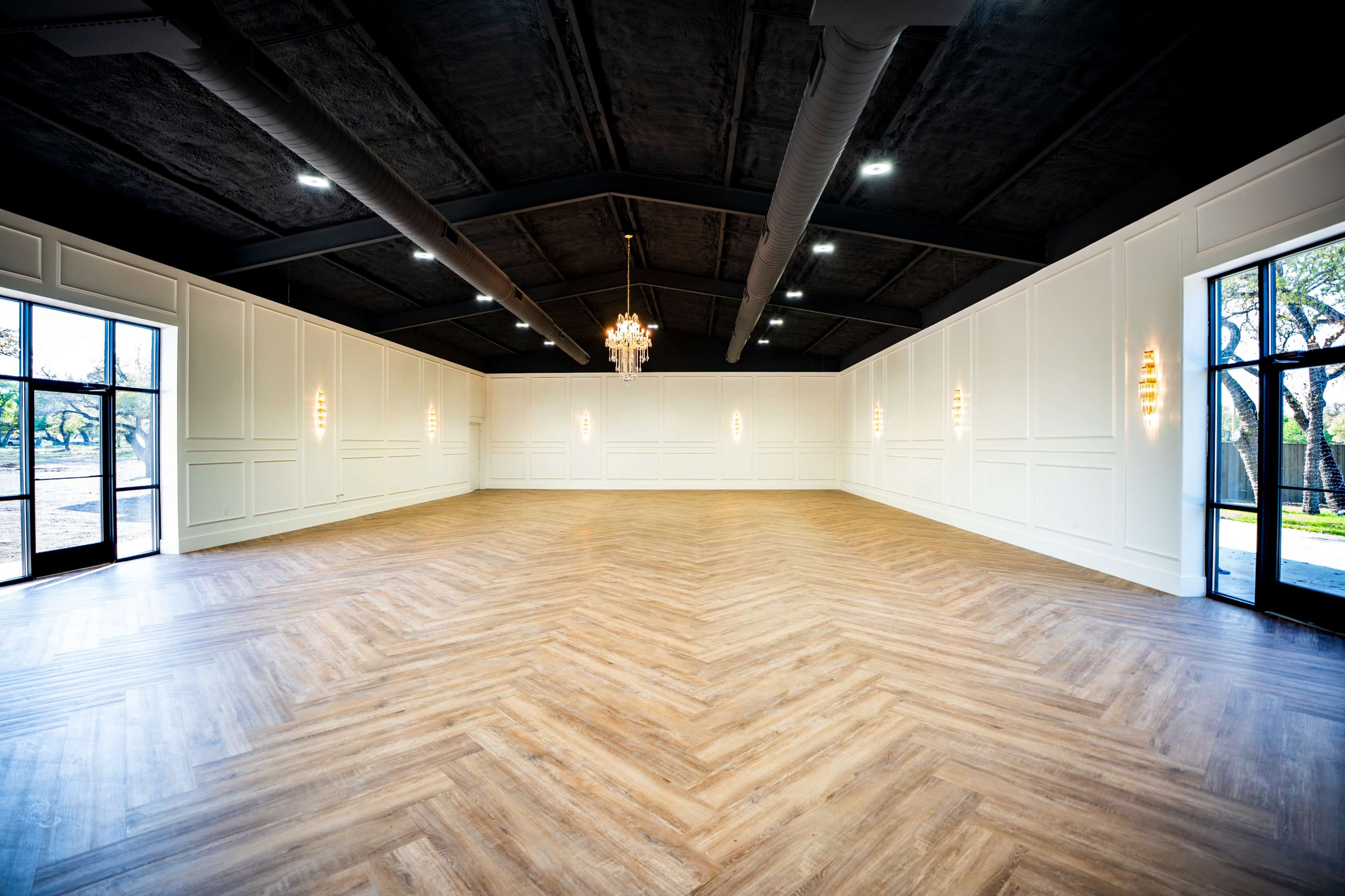 The image shows an empty indoor space with a wooden herringbone floor, white panelled walls, large windows, and a chandelier hanging from the ceiling.