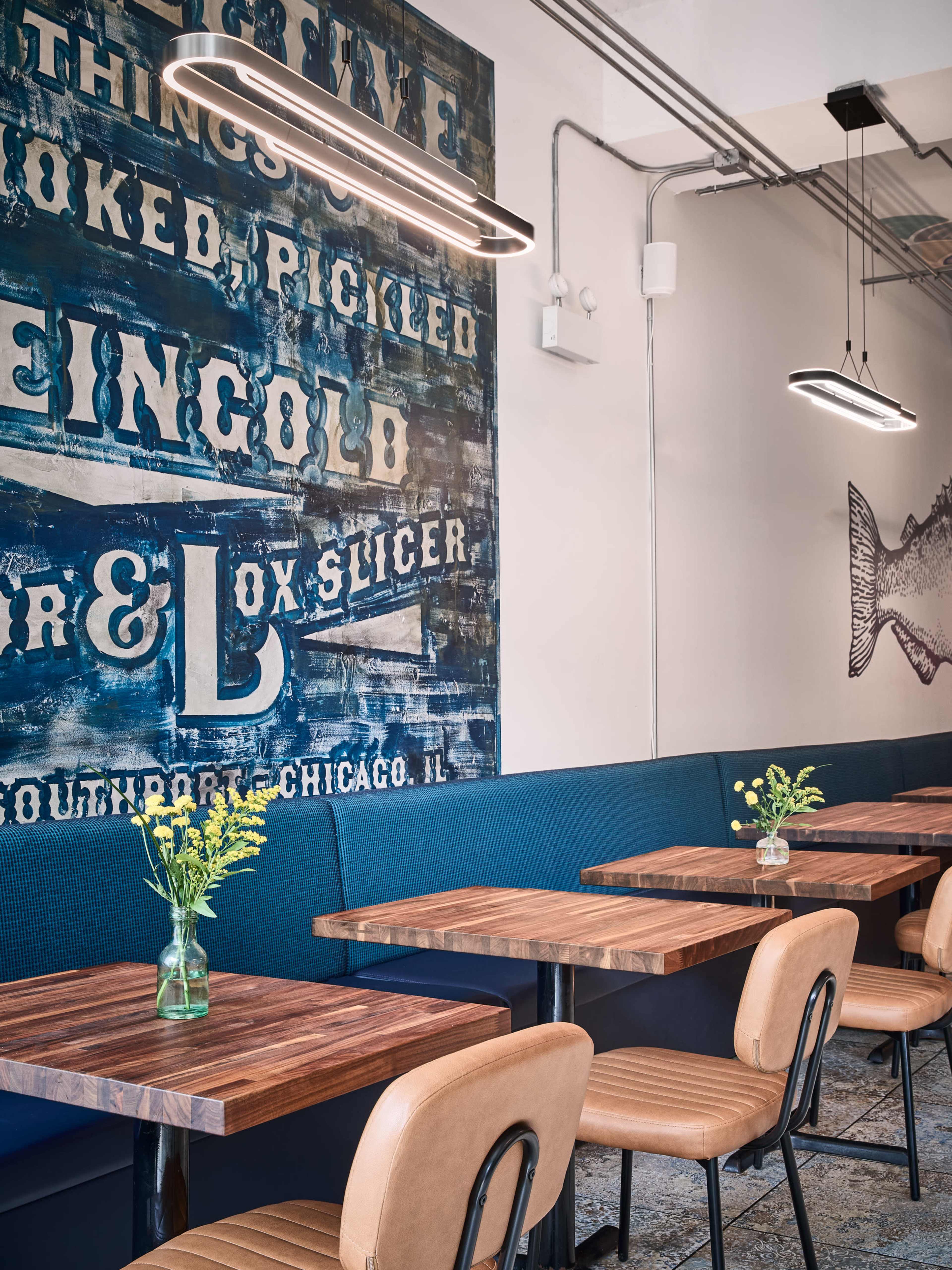 The image shows a modern café interior featuring wooden tables, metal-framed chairs, a blue upholstered wall, and a vintage mural depicting food-related text and a fish.