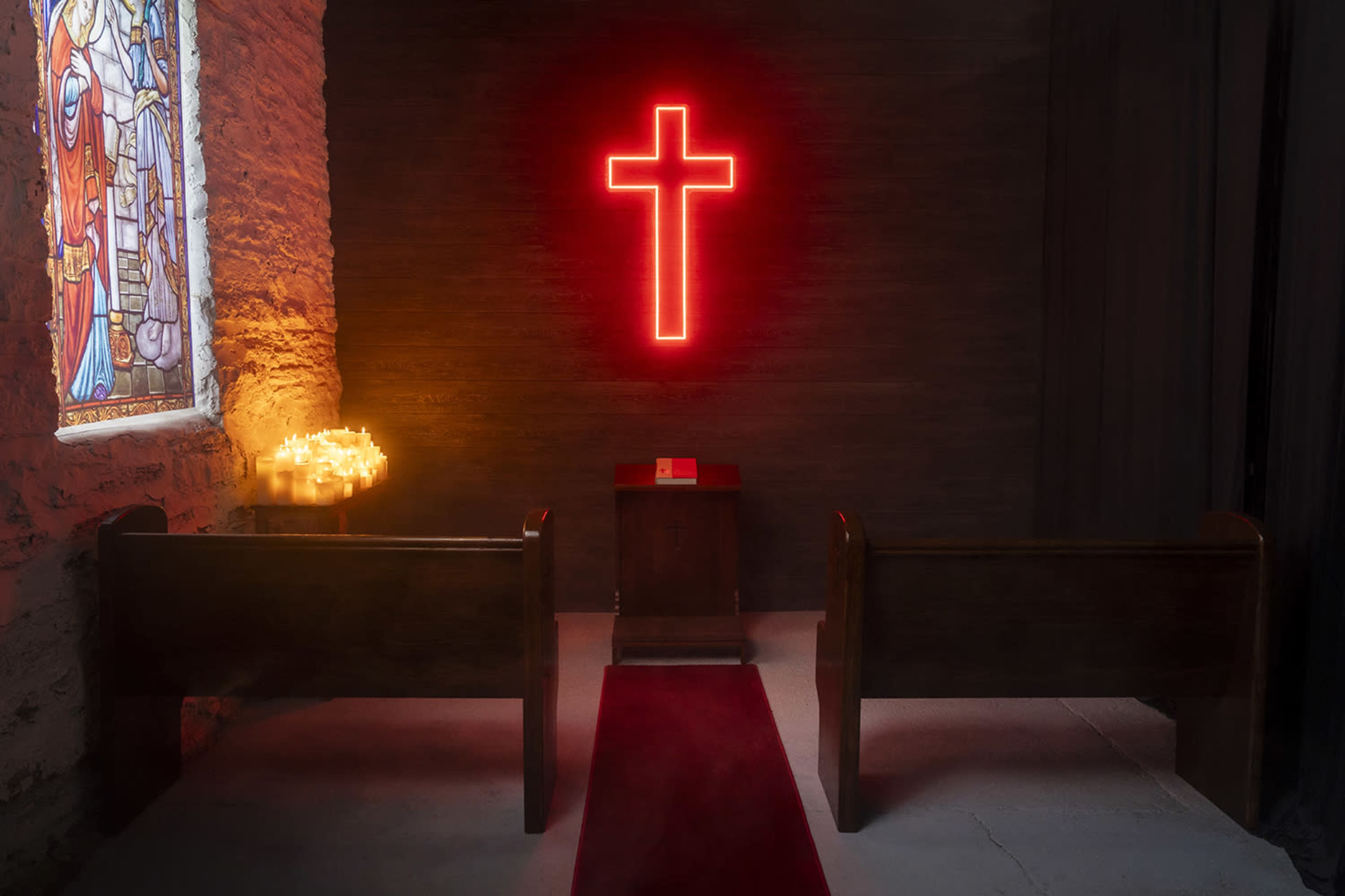 The image shows a dimly lit chapel with wooden pews, a red neon cross on the wall, and a stained glass window.