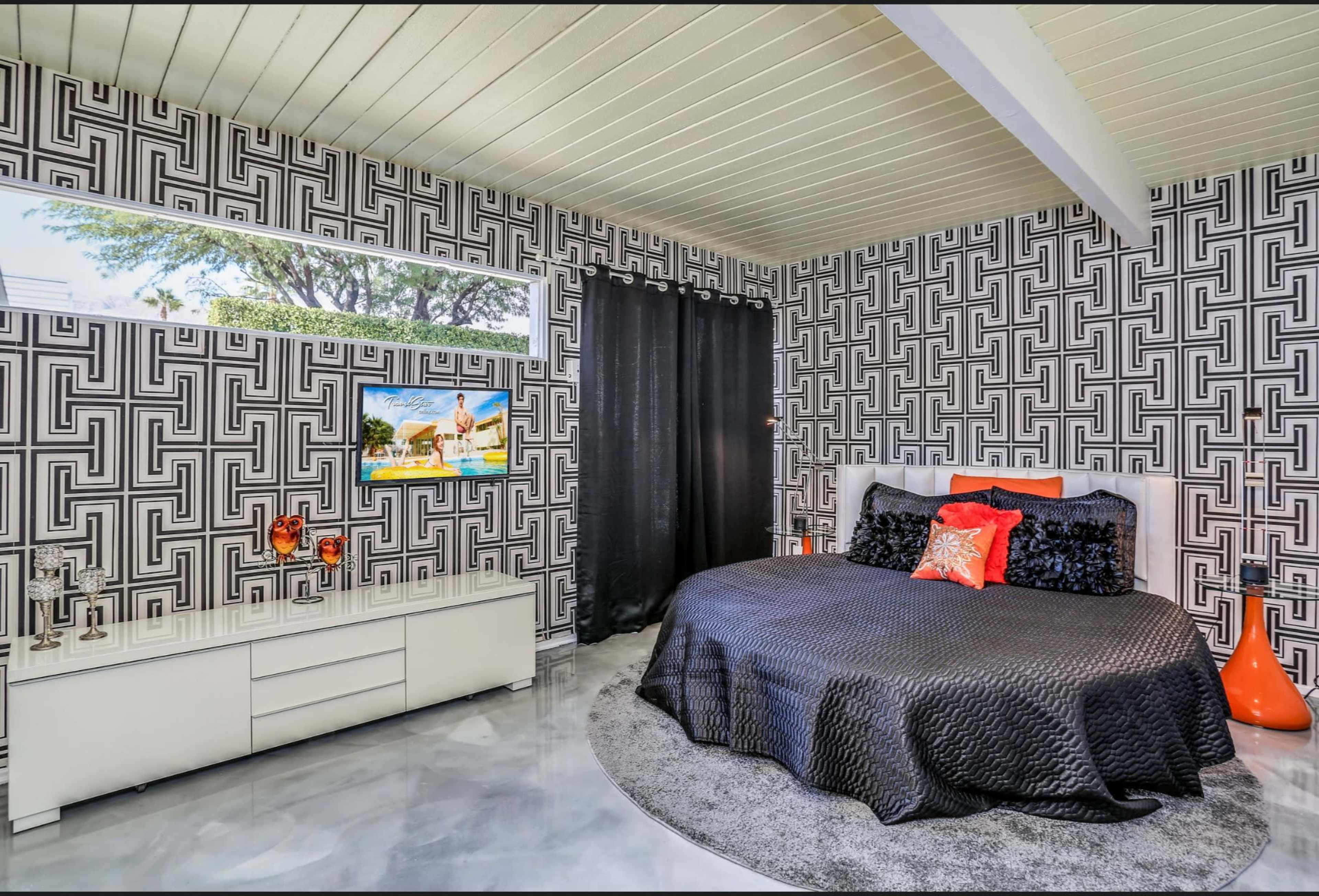 The room features a patterned black and white wallpaper, a large bed with dark bedding, a circular rug, a white dresser, and a large window letting in natural light.
