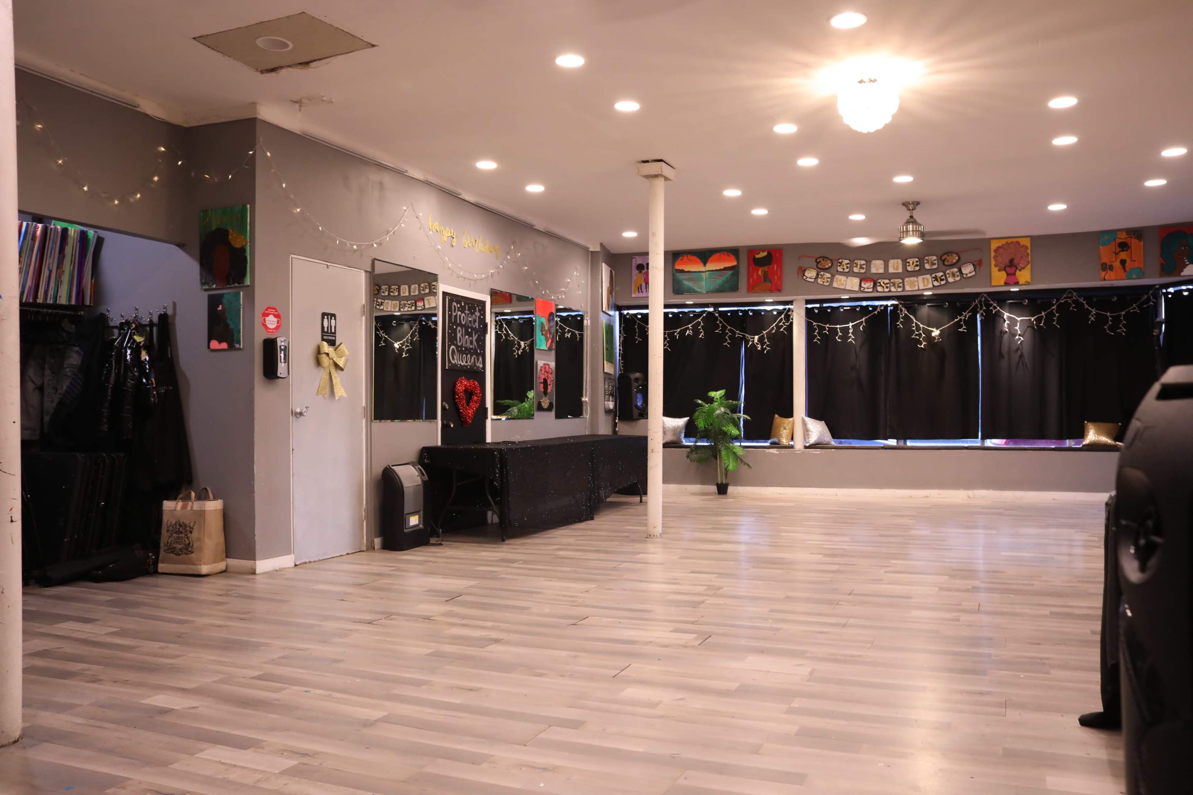 An empty dance studio with gray walls, a polished wooden floor, and mirrors along one side, decorated with artwork and string lights.