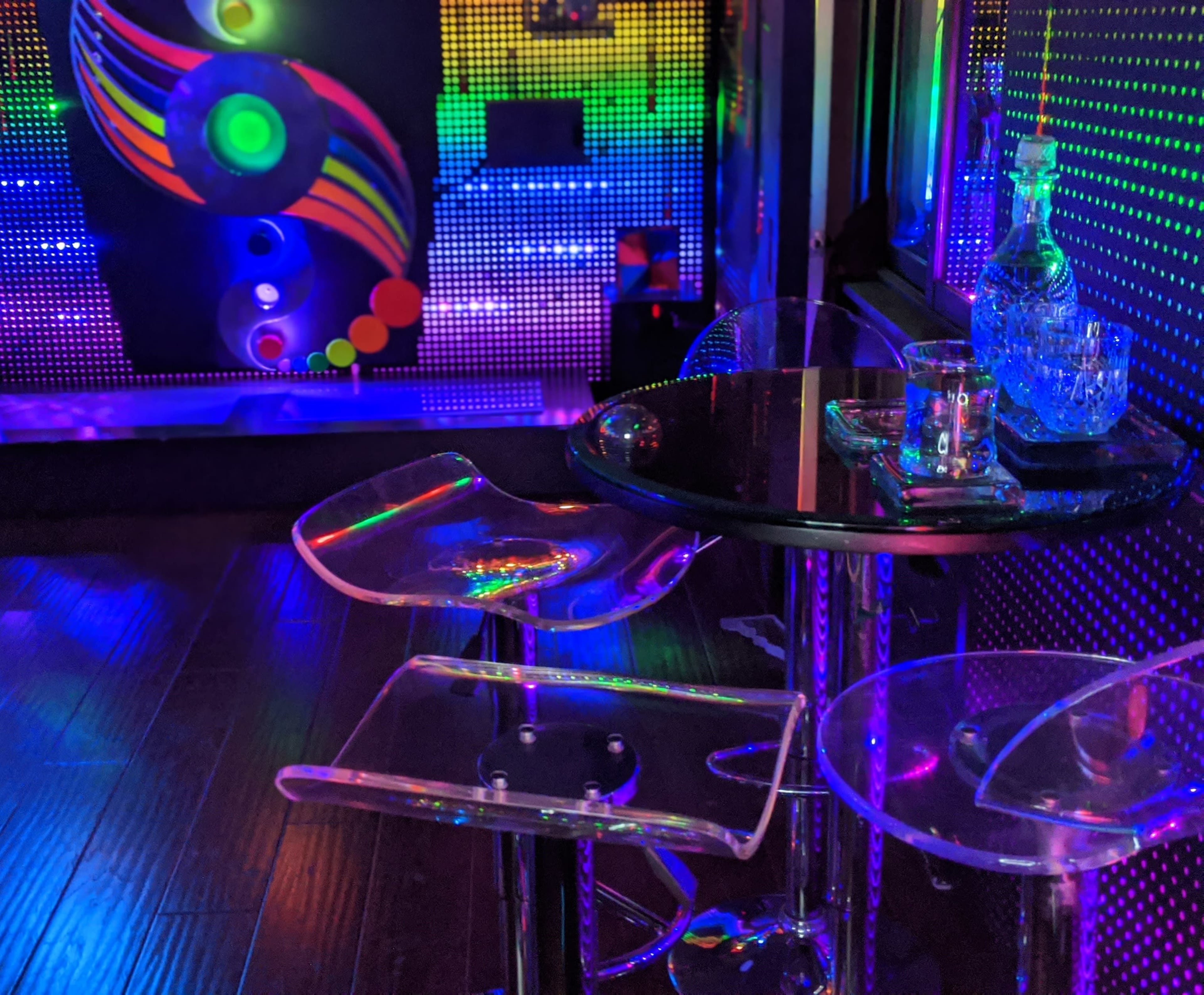The scene features a modern lounge area with colorful LED lights, highlighting transparent furniture and a glass decanter on a black table.