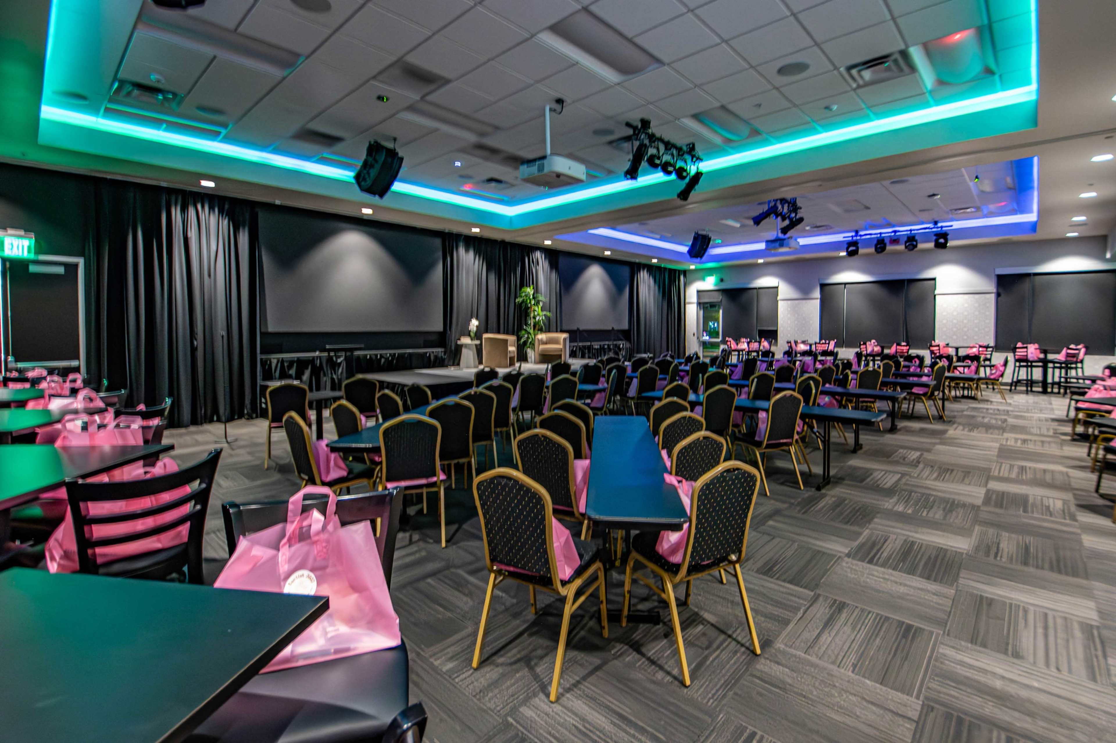 A spacious event room with black chairs and tables, decorated with pink bags on the tables, and equipped with audio-visual equipment and dim lighting.