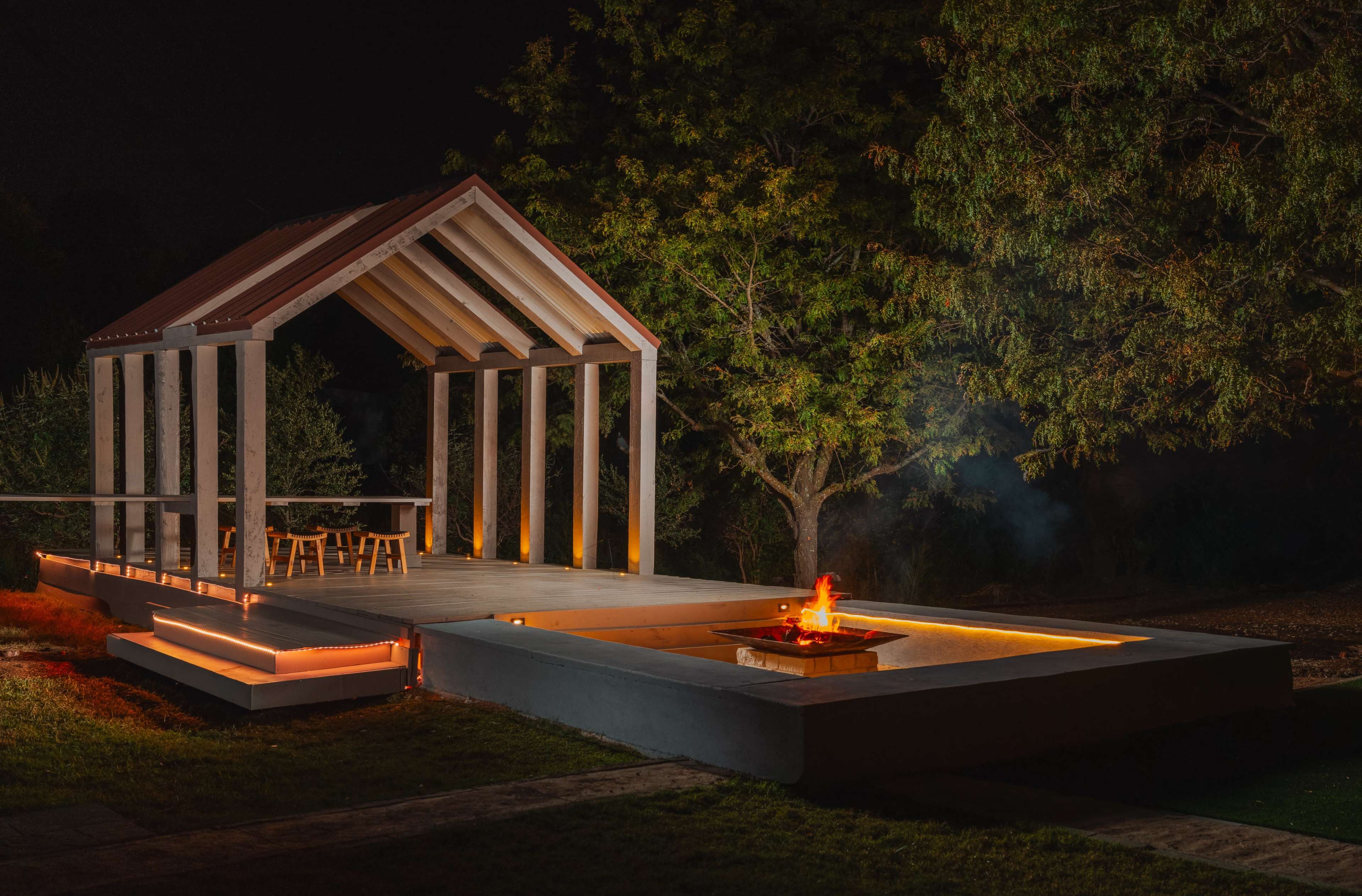 A modern outdoor pavilion with a fire pit is illuminated at night, surrounded by trees and grass.