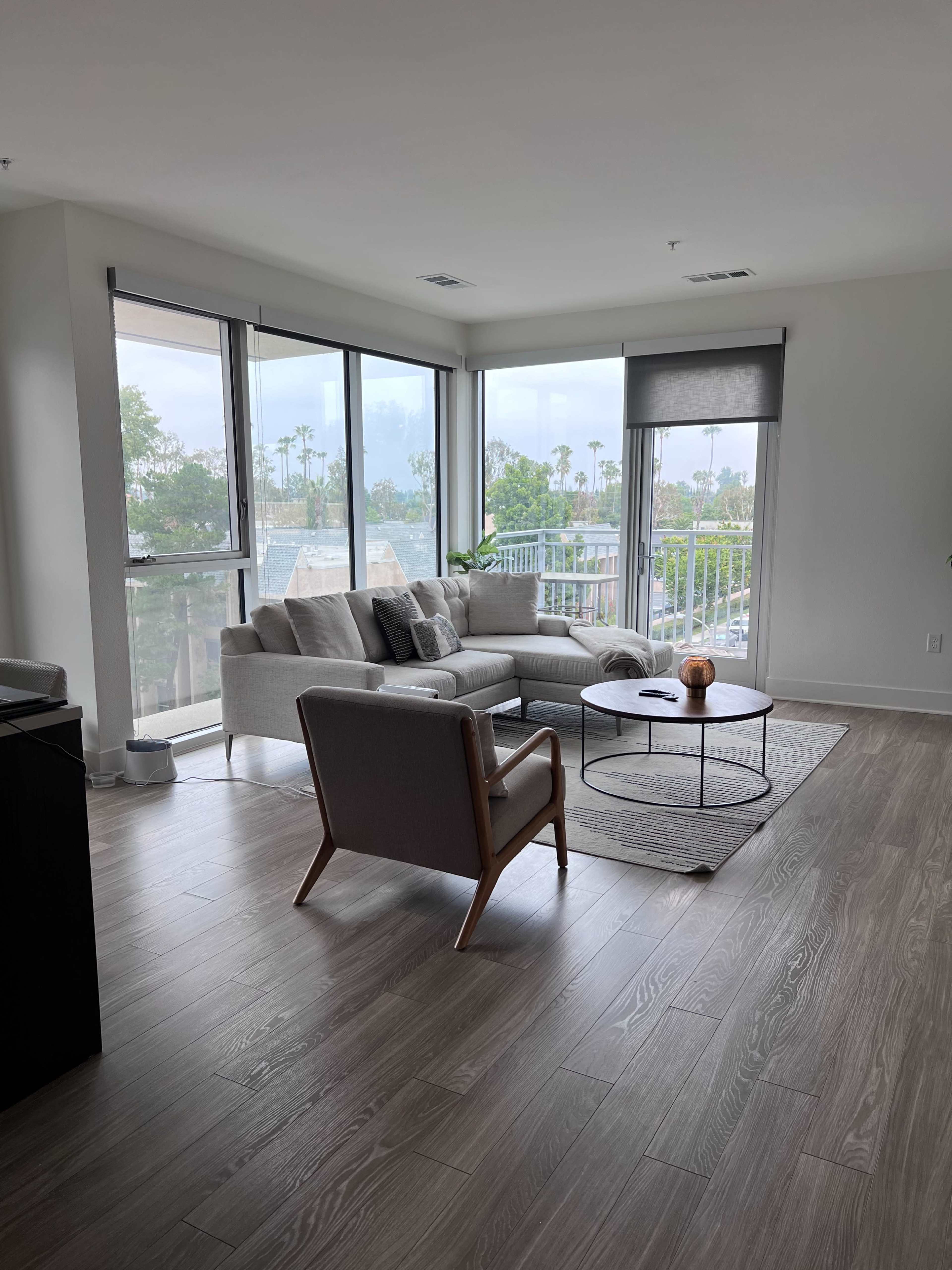 A modern living room features a light gray sofa and an armchair surrounding a circular coffee table, with large windows offering views of palm trees outside.