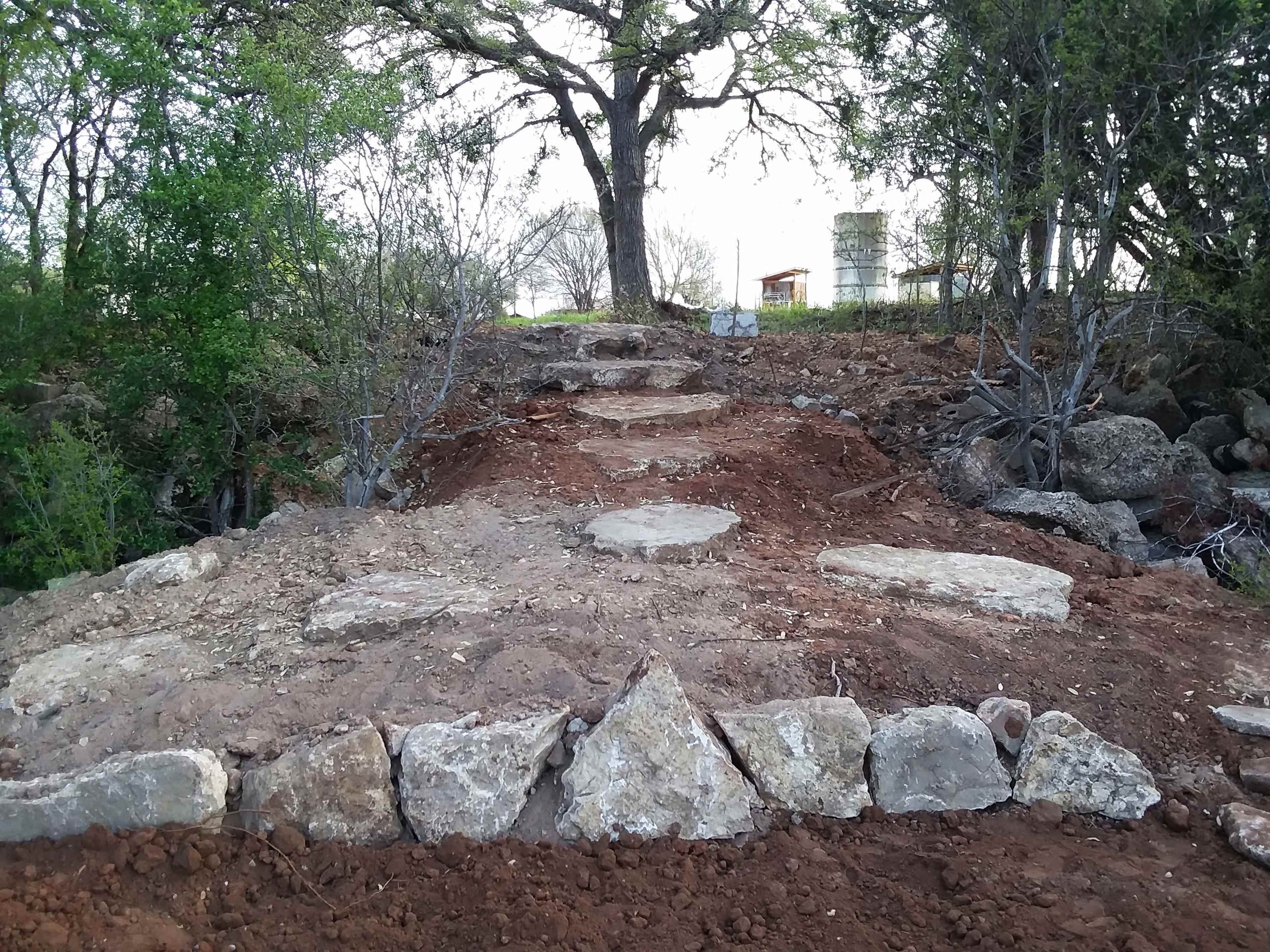 A path made of large stone slabs leads up a dirt incline, flanked by sparse vegetation and a tree in the background.