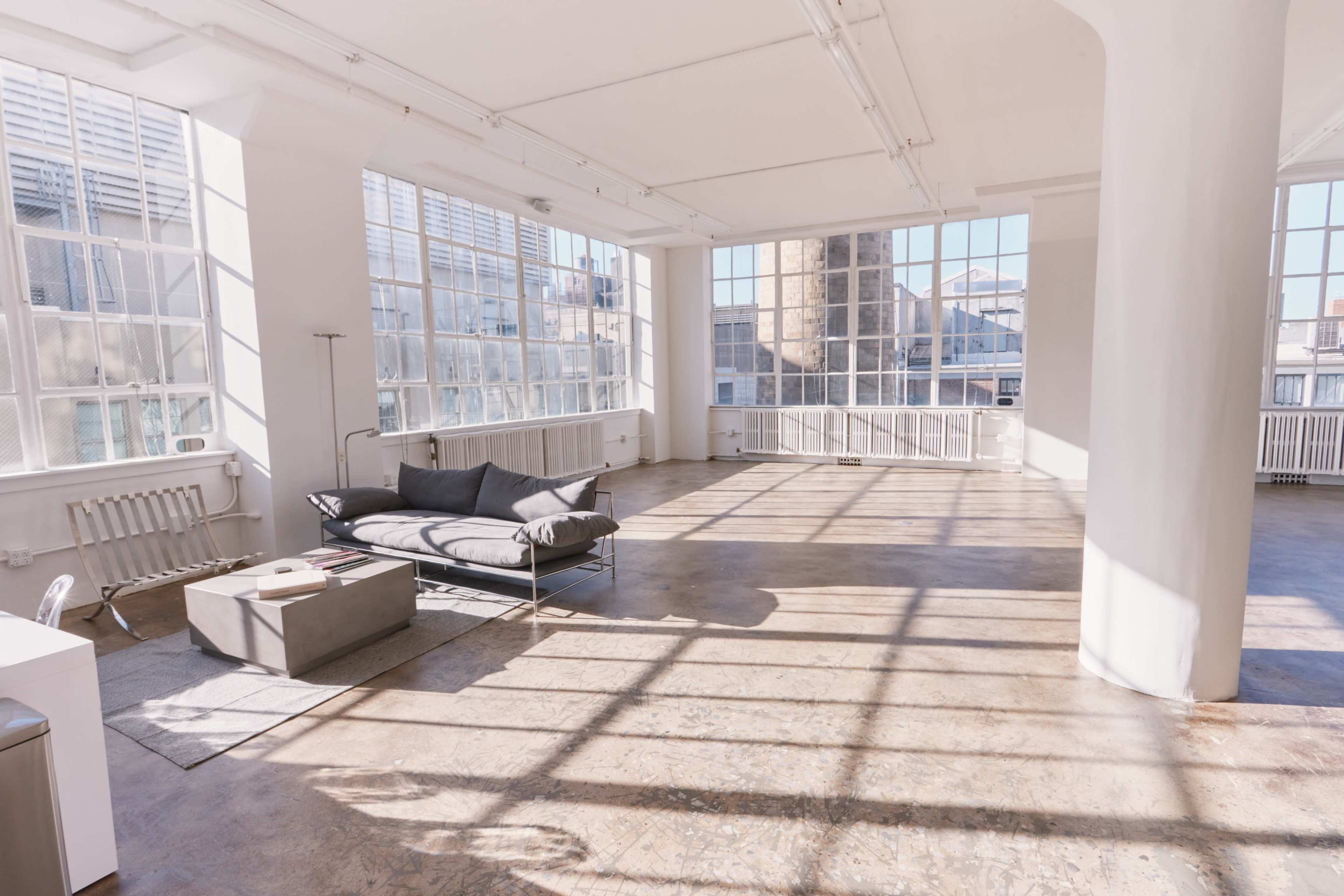 A spacious, well-lit room with large windows, a gray sofa, and minimalist furniture.