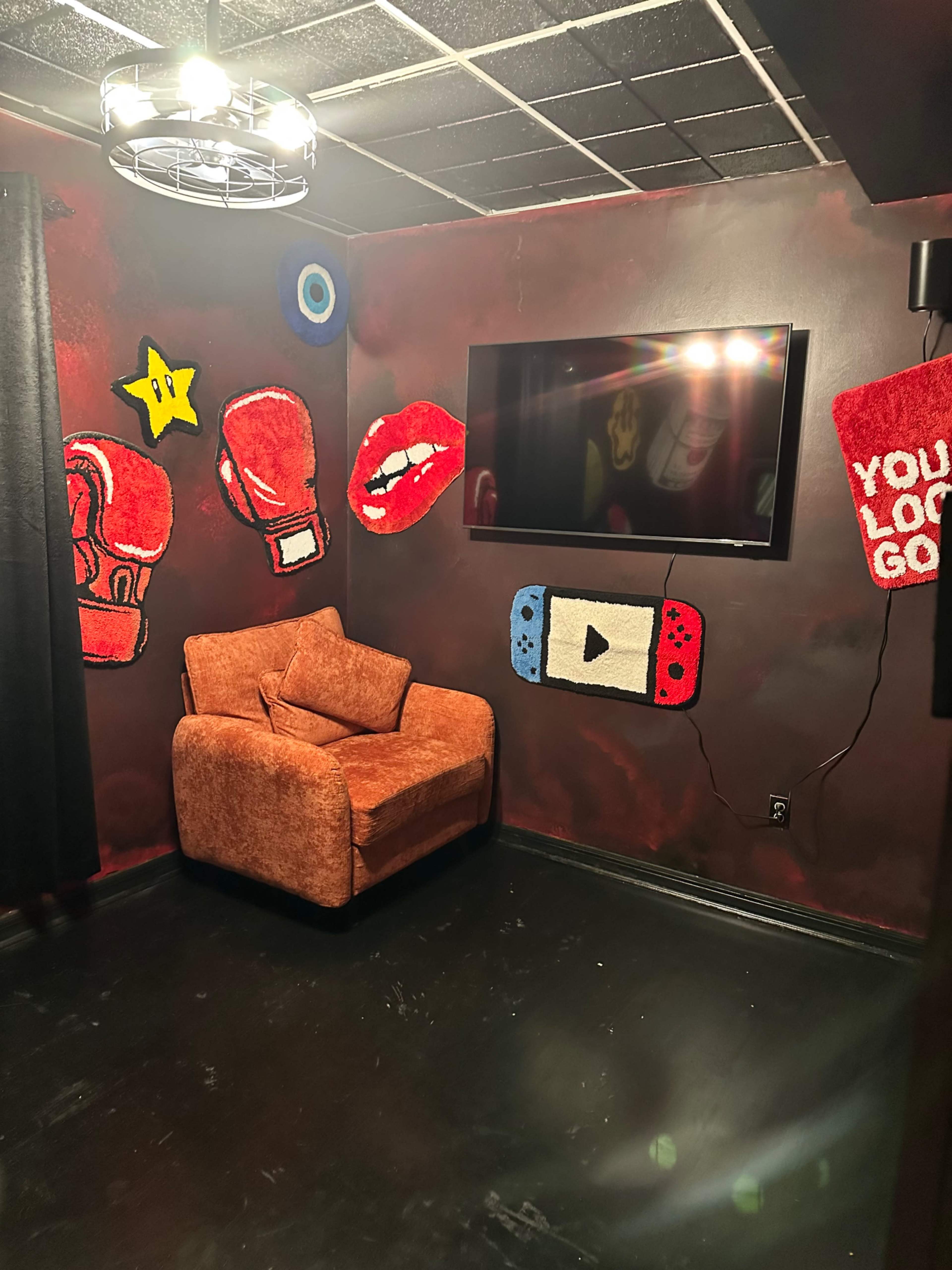 The image shows a small room with a brown armchair, a flat-screen TV mounted on the wall, and colorful decorations, including cartoonish boxing gloves, lips, a star, and a Nintendo Switch design on the walls.