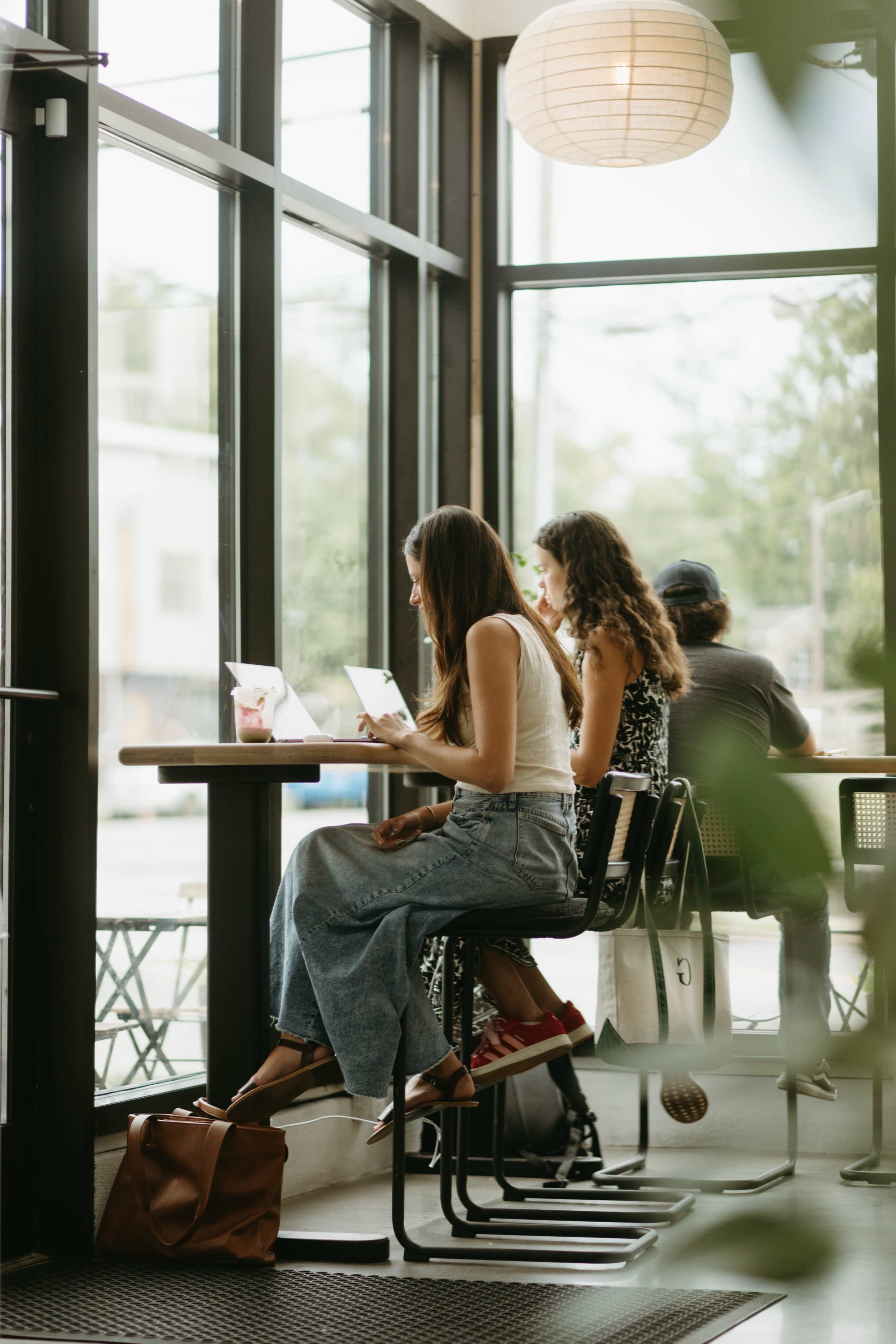 Two women are sitting at a café table, working on laptops while a man sits nearby, with bright natural light coming through large windows.