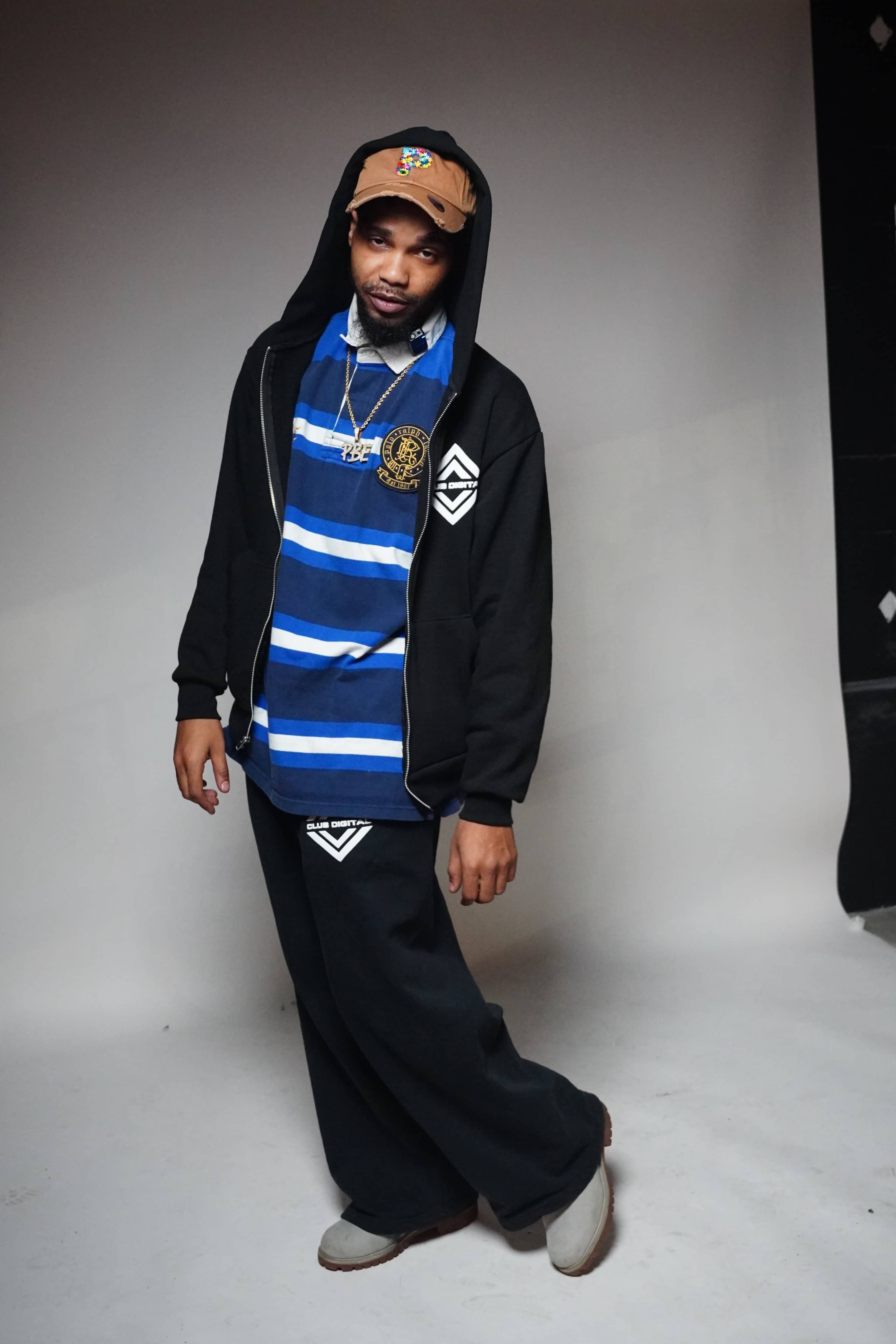 A person stands in a studio wearing a blue and white striped shirt, black hoodie, and loose black pants, with a cap and necklace.