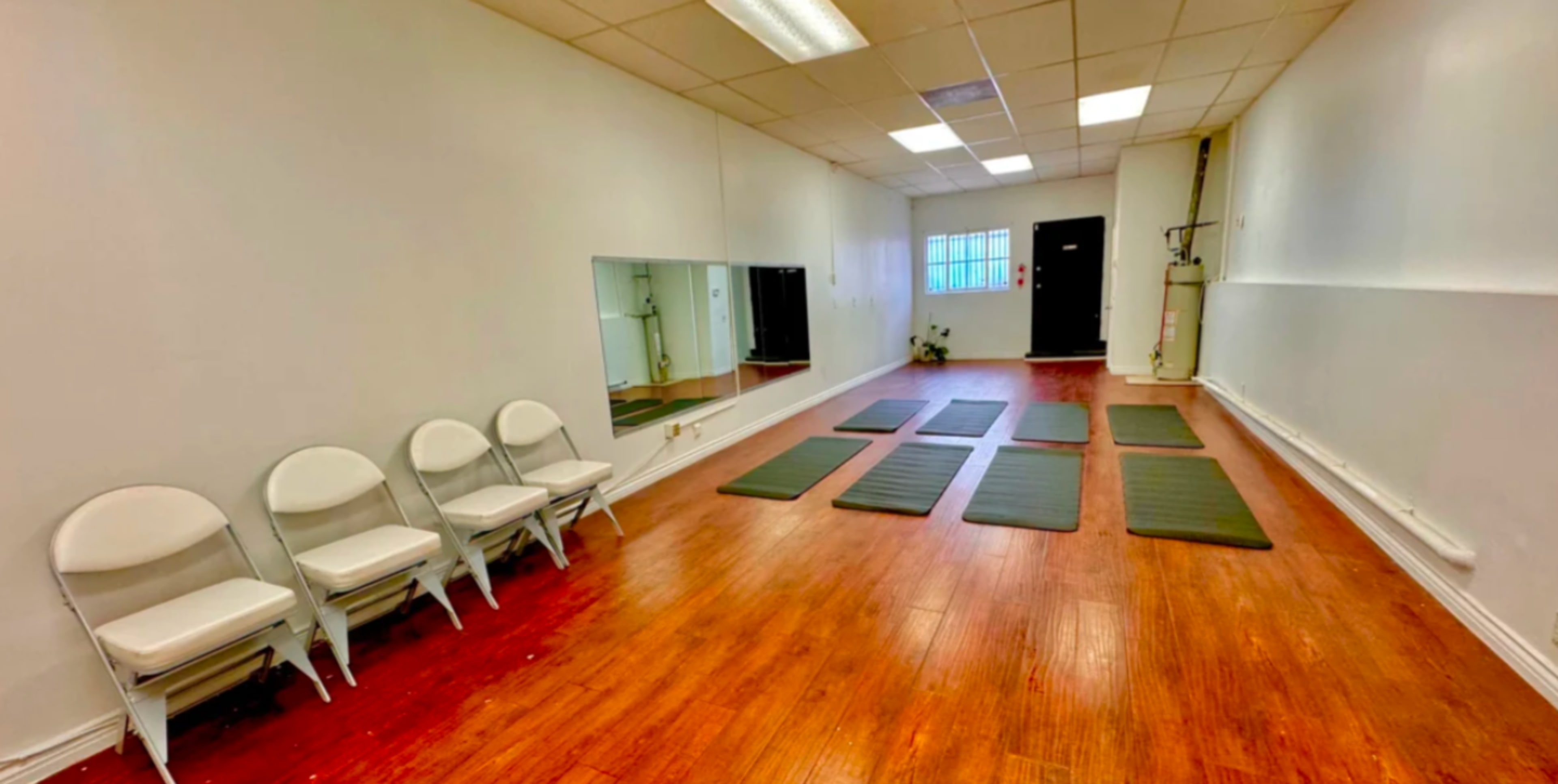 A spacious, minimalist room with wooden flooring, four exercise mats on the floor, and a row of white chairs against one wall.