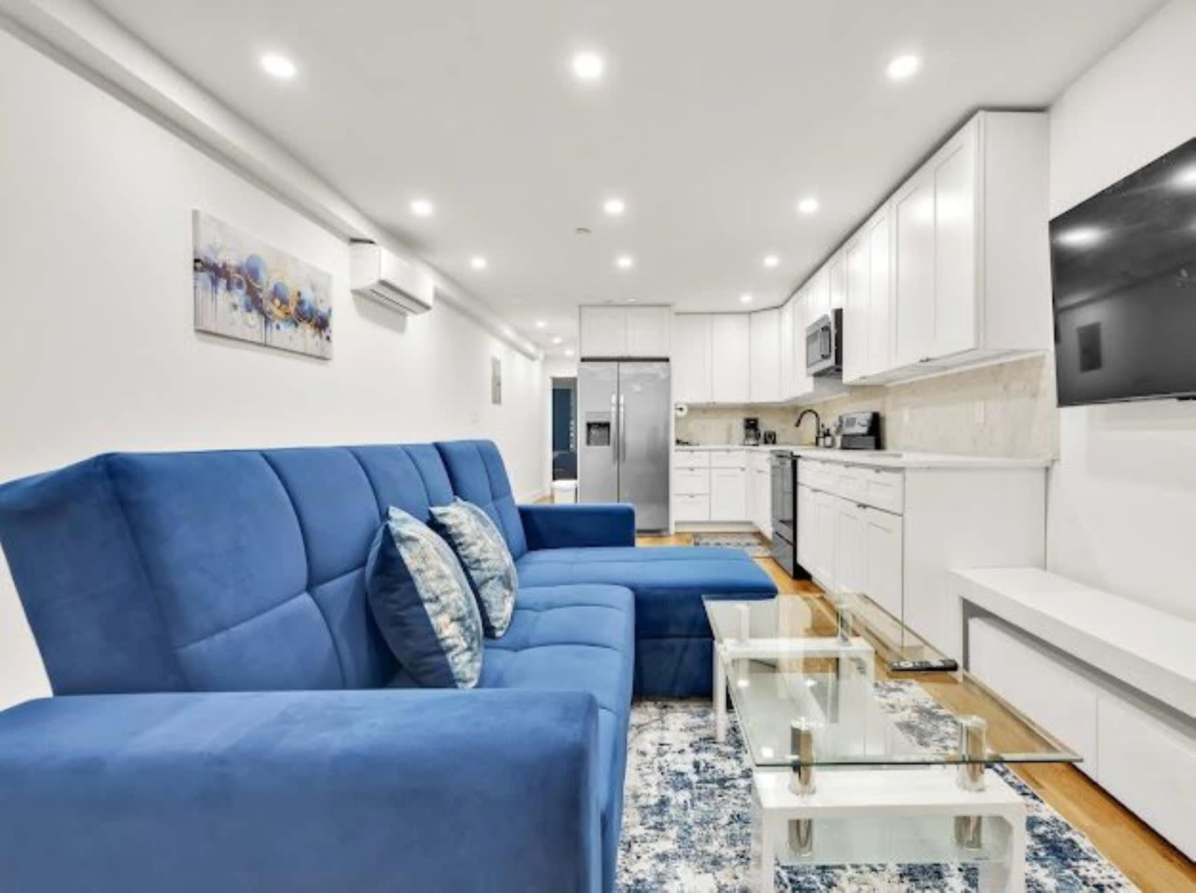 This modern living space features a blue sectional sofa, a glass coffee table, and a kitchen with white cabinetry and stainless steel appliances.