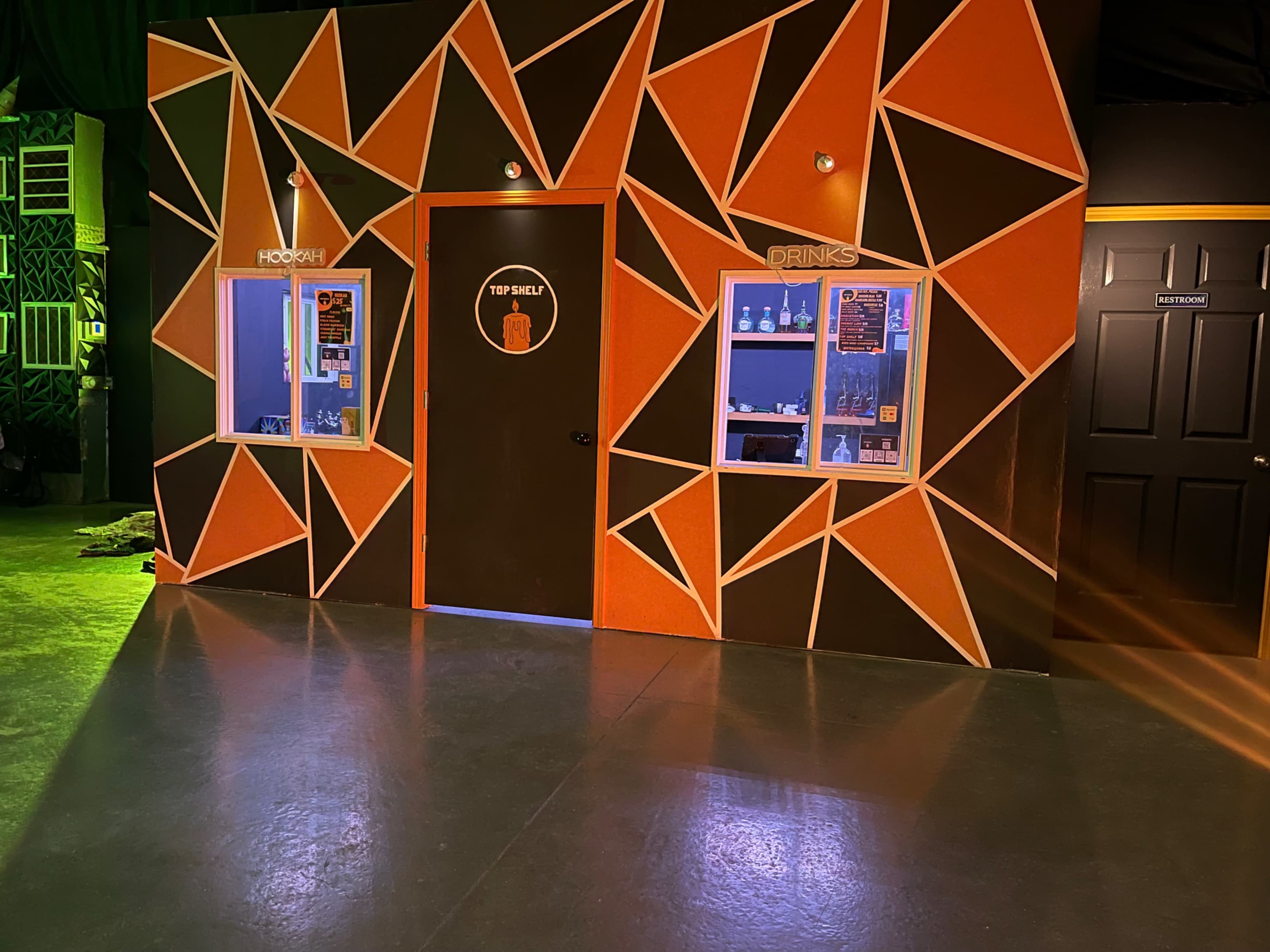 A vibrantly designed wall featuring geometric patterns in orange and black, with windows labeled "HOOCH" and "DRINKS," and a door in the center labeled "TOP SHELF."
