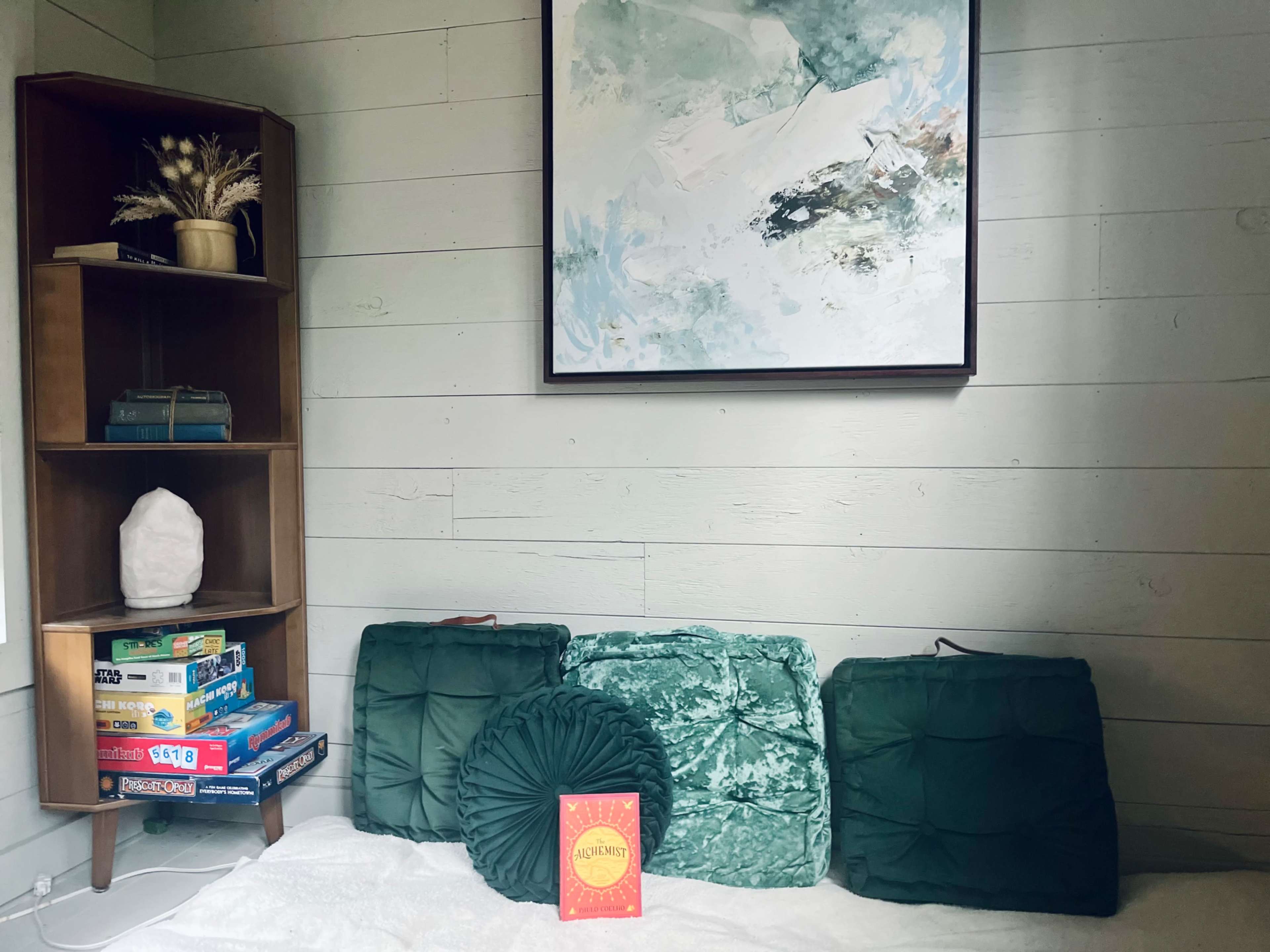 A cozy interior featuring a shelf with books, a decorative piece, and three green cushions on a white surface, with an art piece hanging on the wall.