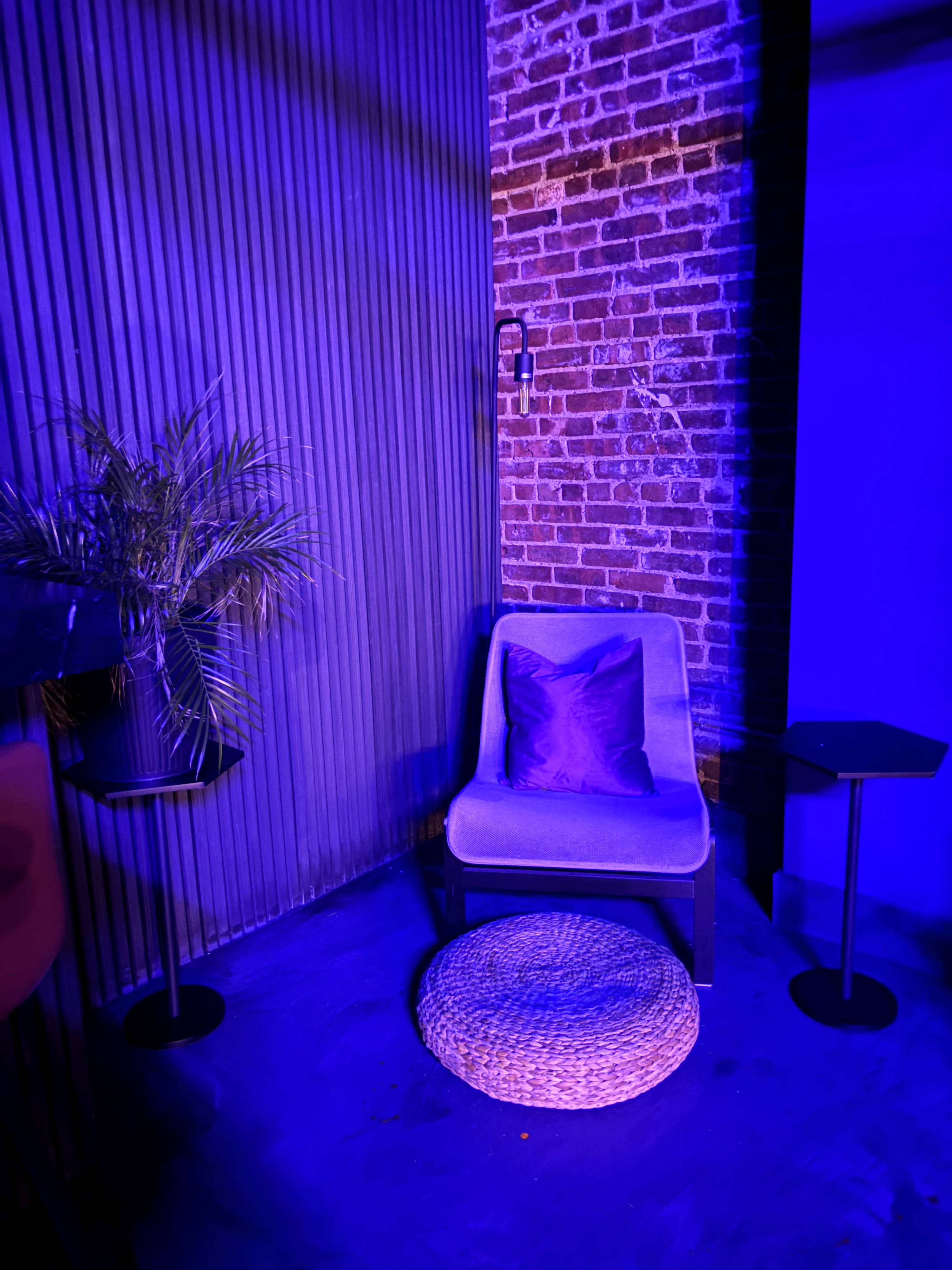 The image shows a seating area with a gray chair, a round woven pouf, a small side table, and a potted plant, all illuminated by a purple light against a backdrop of exposed brick walls.