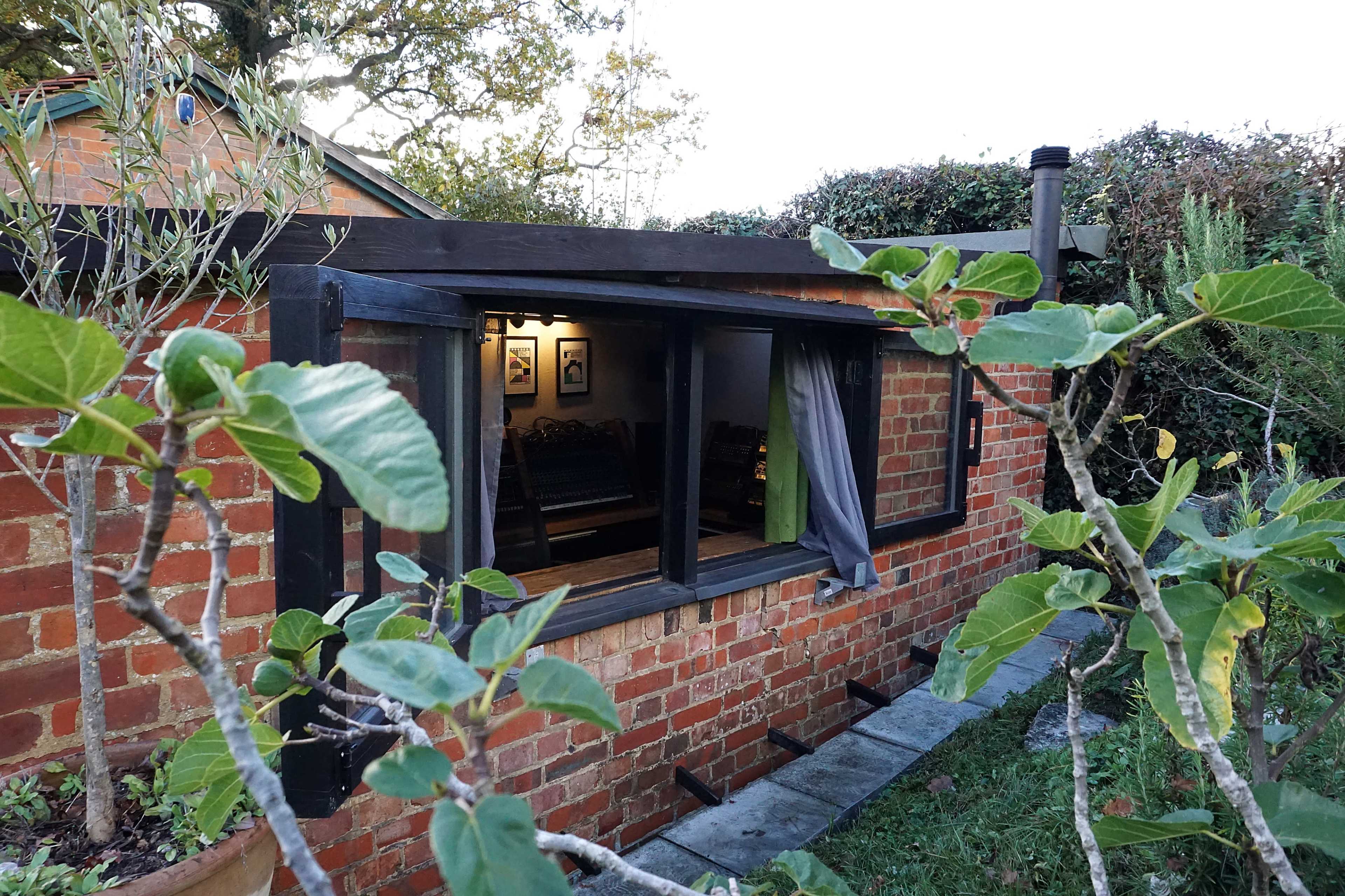 A brick building with a large window is surrounded by greenery, featuring a visible interior space with furniture.