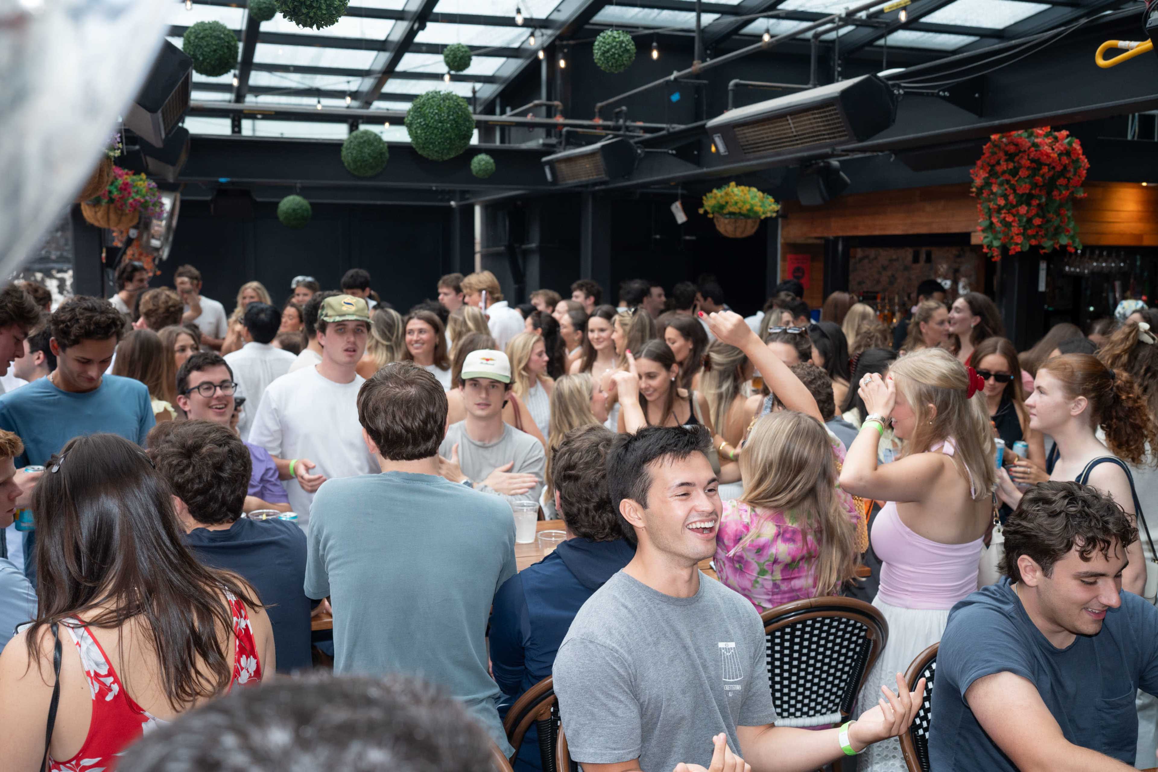A lively indoor party scene shows a large group of young adults socializing at tables with drinks and food, surrounded by decorative hanging plants.
