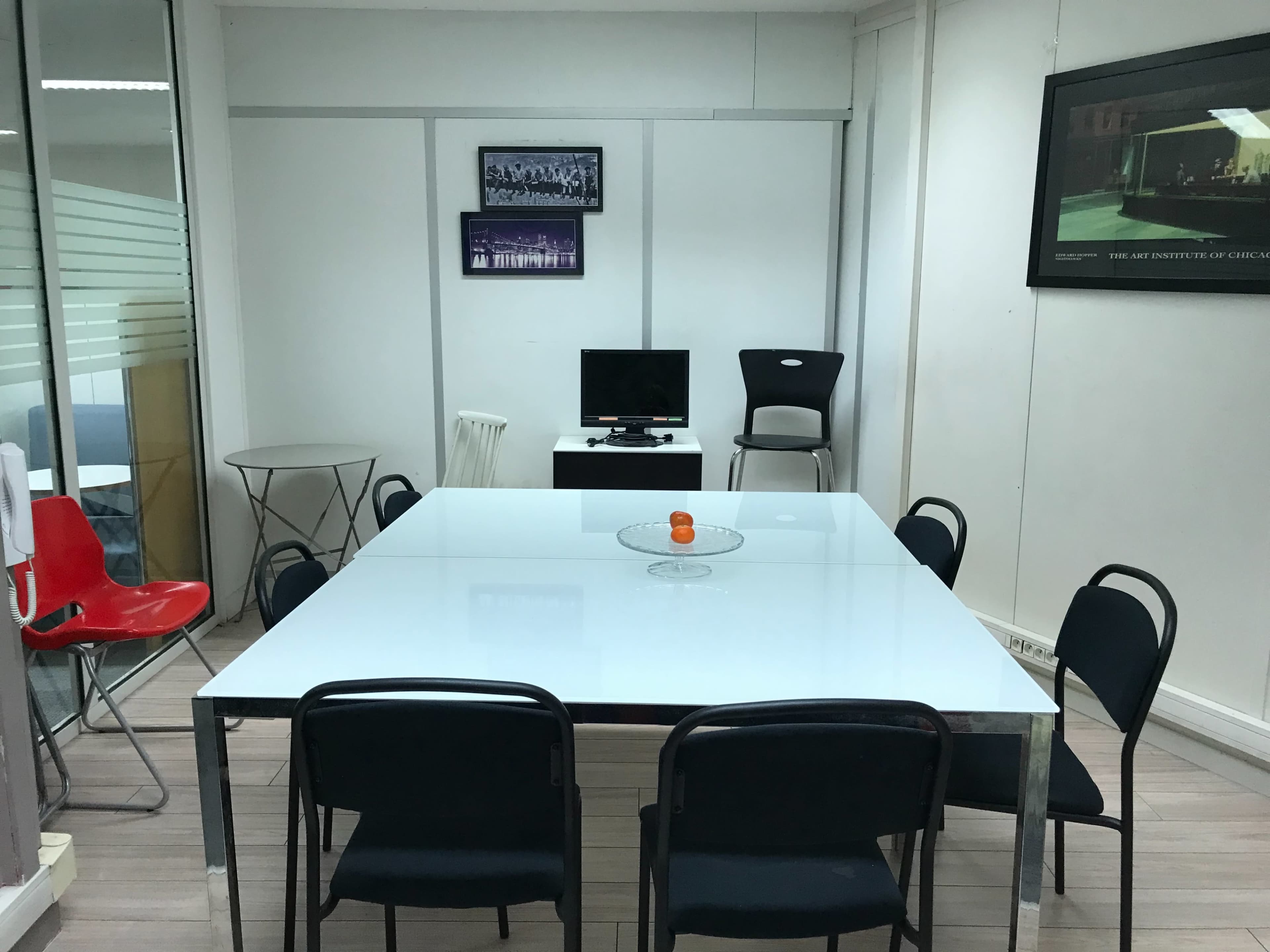A conference room features a large white table surrounded by black chairs, with a red chair, a television, and framed artworks on the walls.