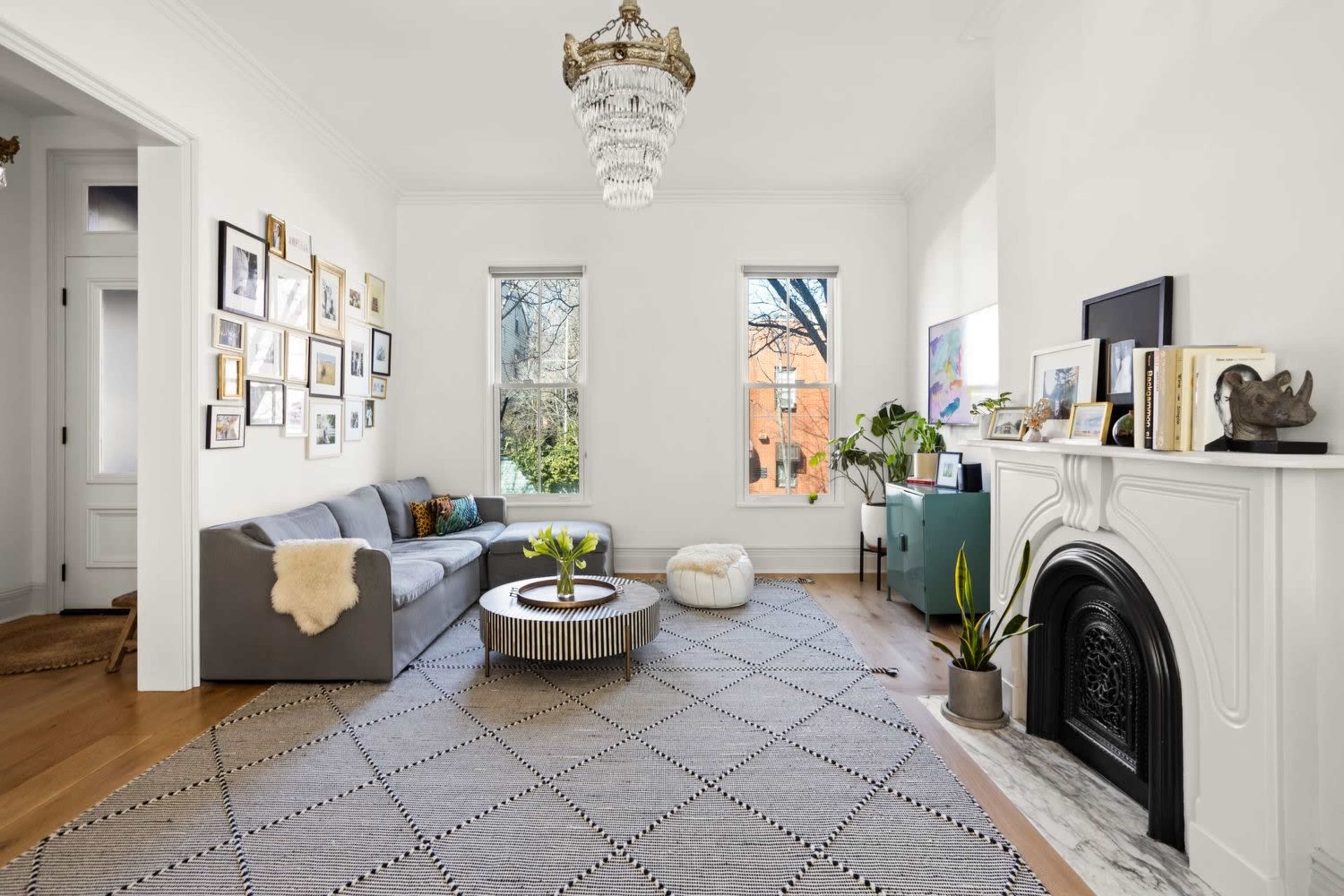 The image shows a bright living room with a gray sectional sofa, a round coffee table, and a decorative fireplace, adorned with framed photos and plants.