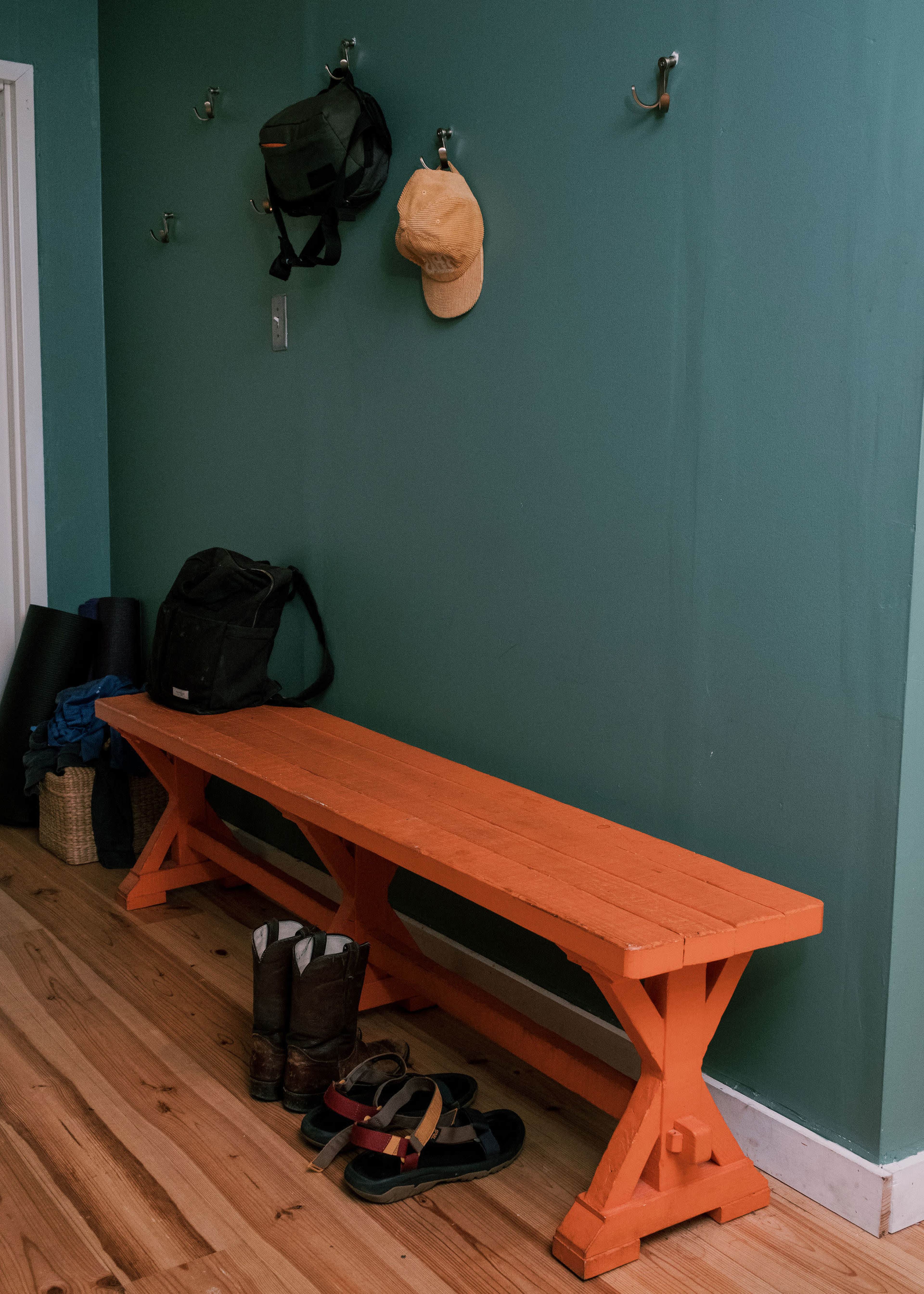The image shows an orange bench against a green wall, with hooks above holding a hat and bags, alongside a pair of boots and sandals on the floor.