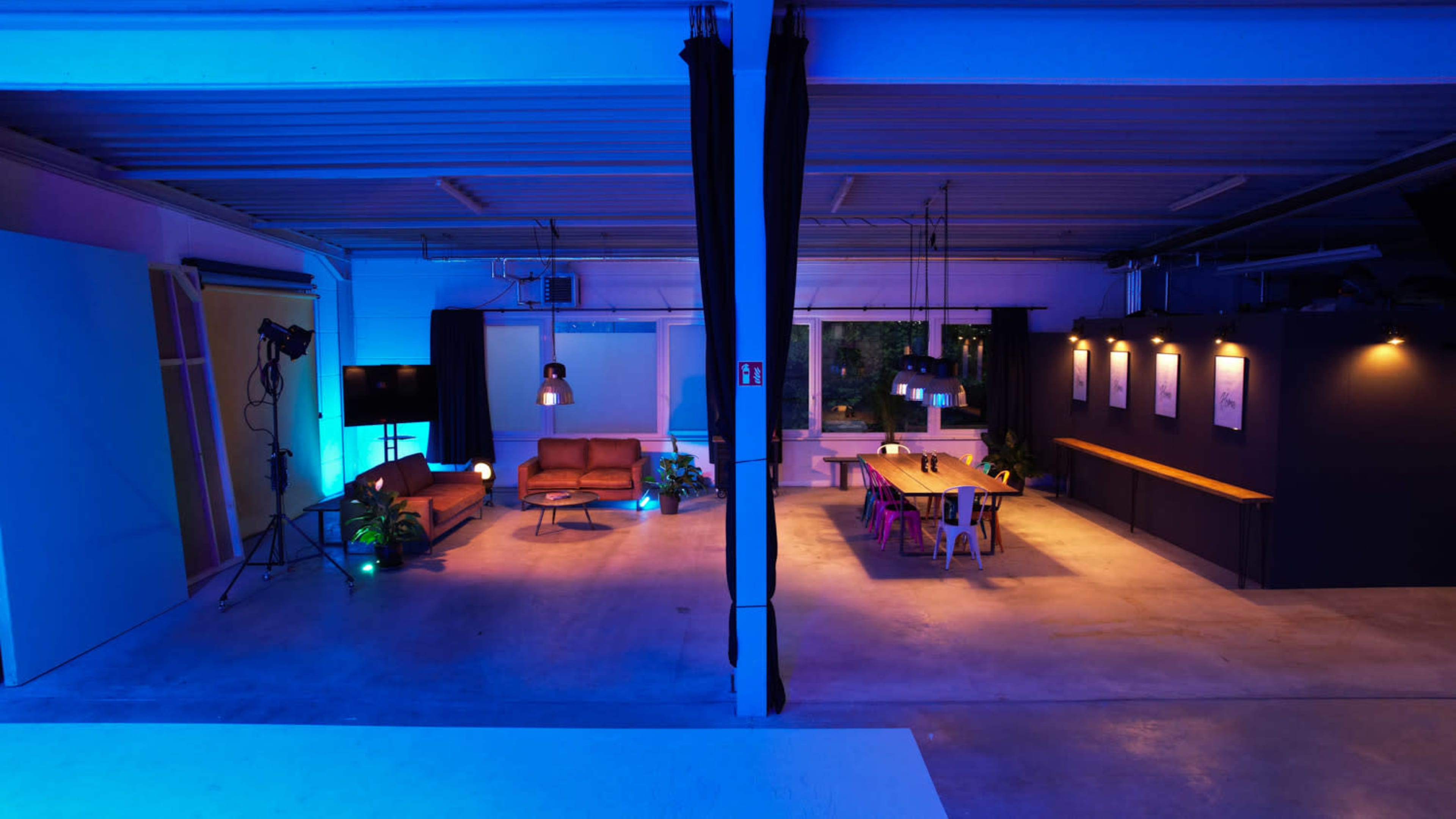 The image shows a modern space divided into two areas: one with a low sofa and a lamp, and the other featuring a long table with chairs under soft lighting.