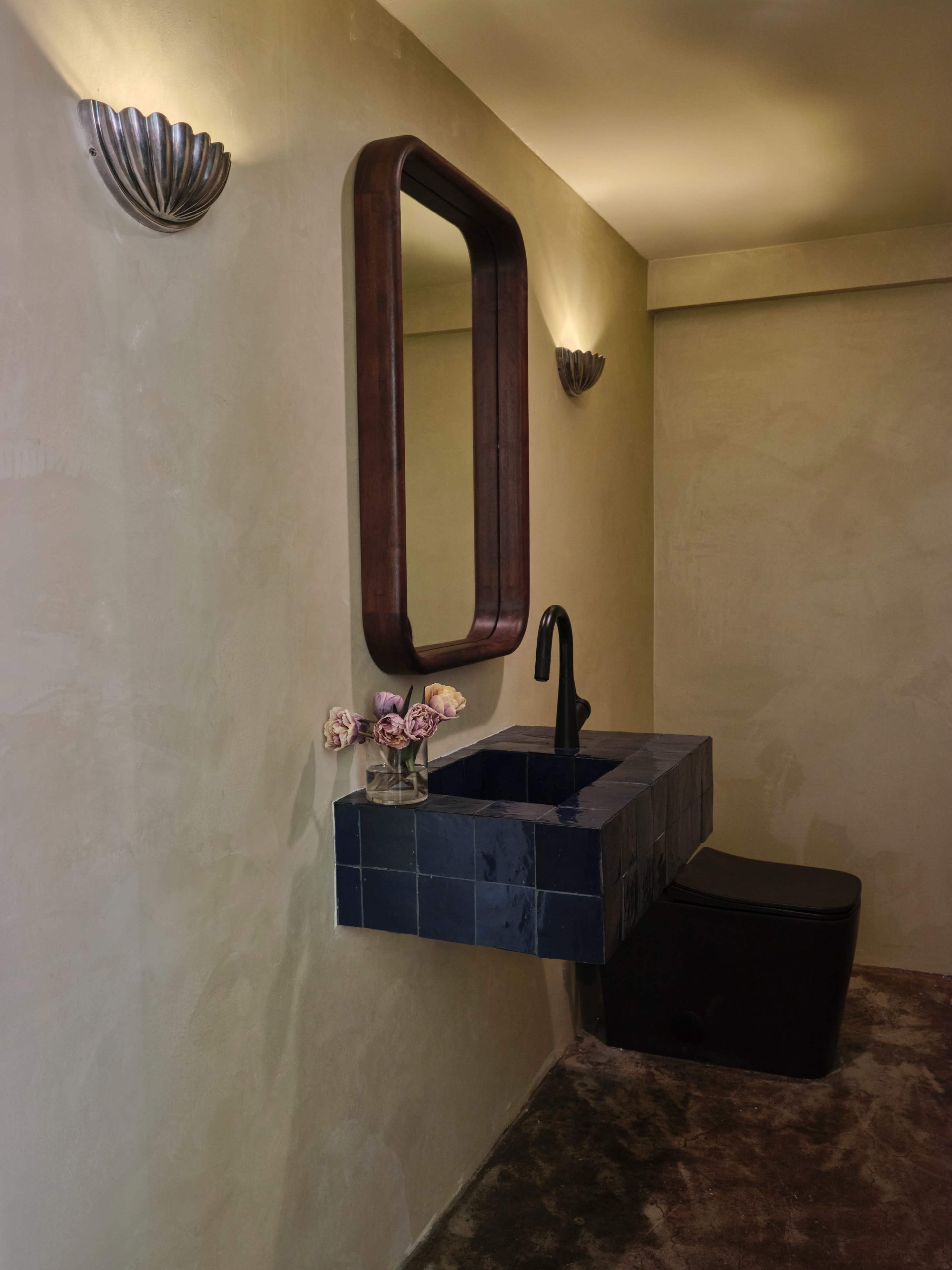 The image shows a minimalistic bathroom featuring a wall-mounted sink with a dark countertop, a large wooden-framed mirror, and a black toilet, all set against a textured beige wall with decorative wall sconces.
