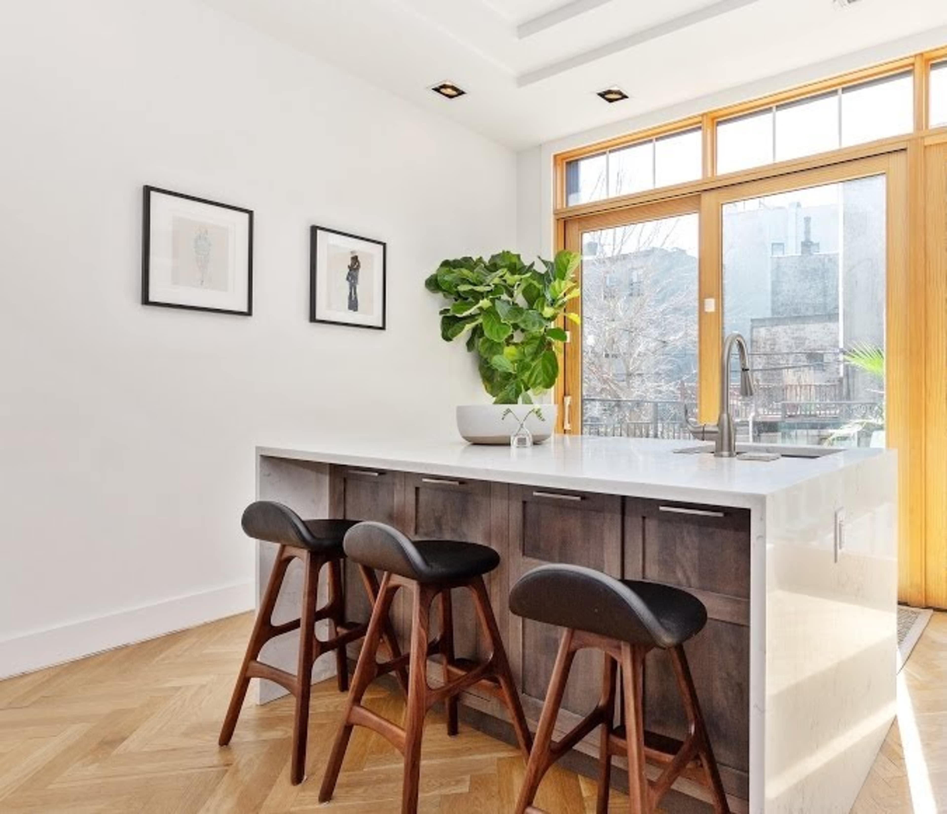 A modern kitchen features a central island with three wooden stools and large windows that let in natural light.