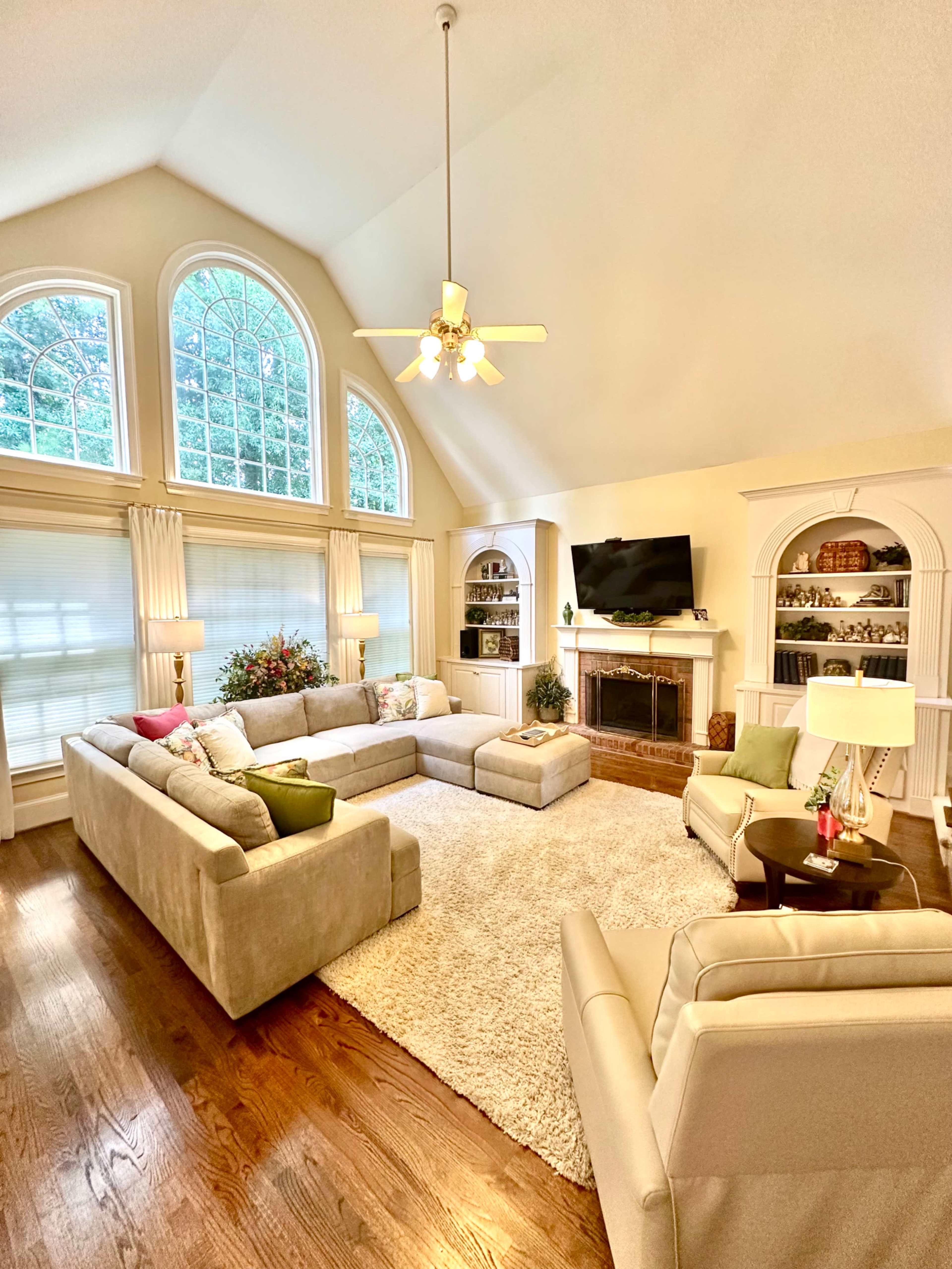 The image shows a spacious living room with large arched windows, a plush sectional sofa, and a fireplace surrounded by built-in shelves.
