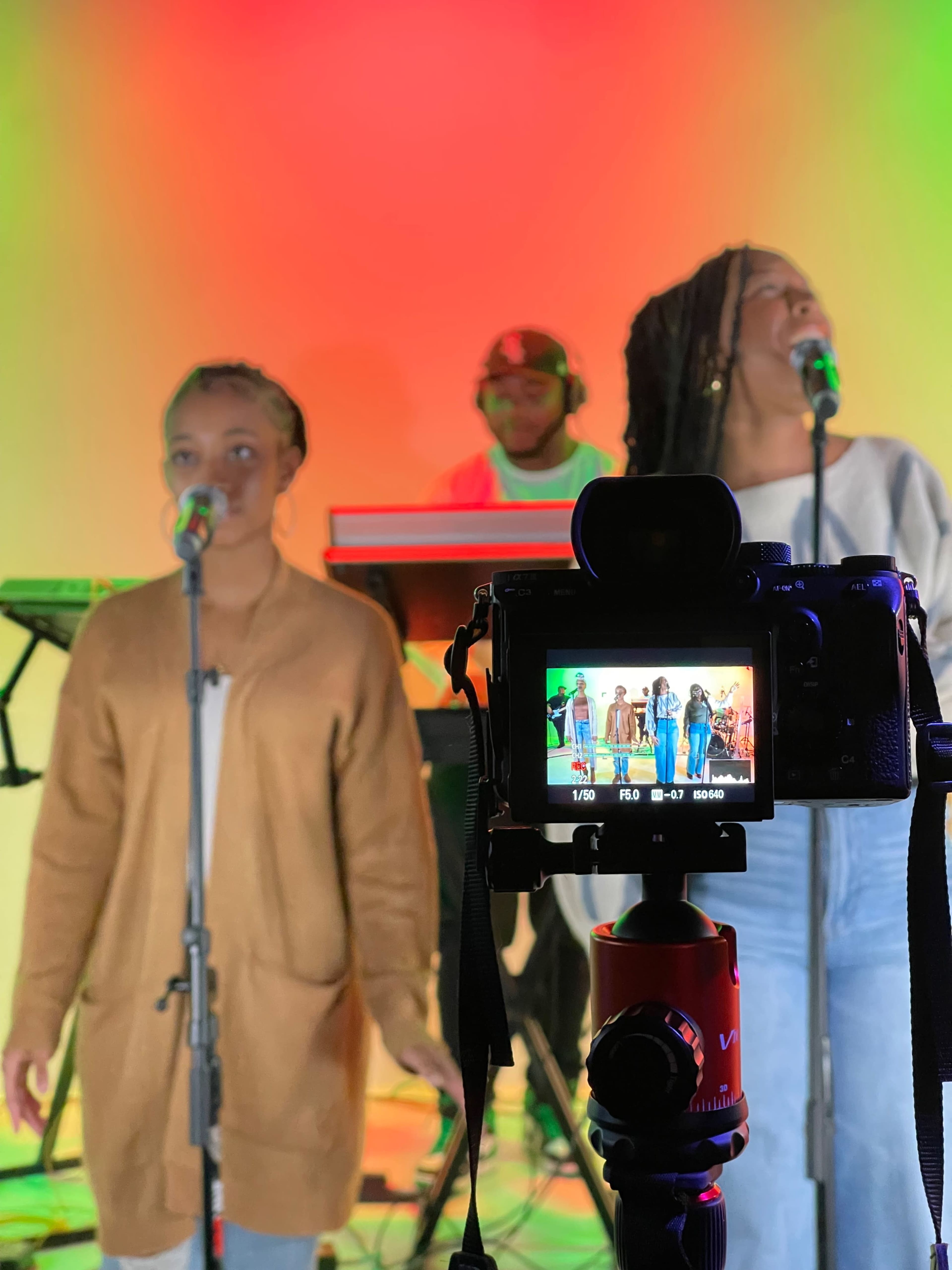 Two singers perform in front of a colorful backdrop while a musician plays a keyboard in the background, with a camera focused on the scene.