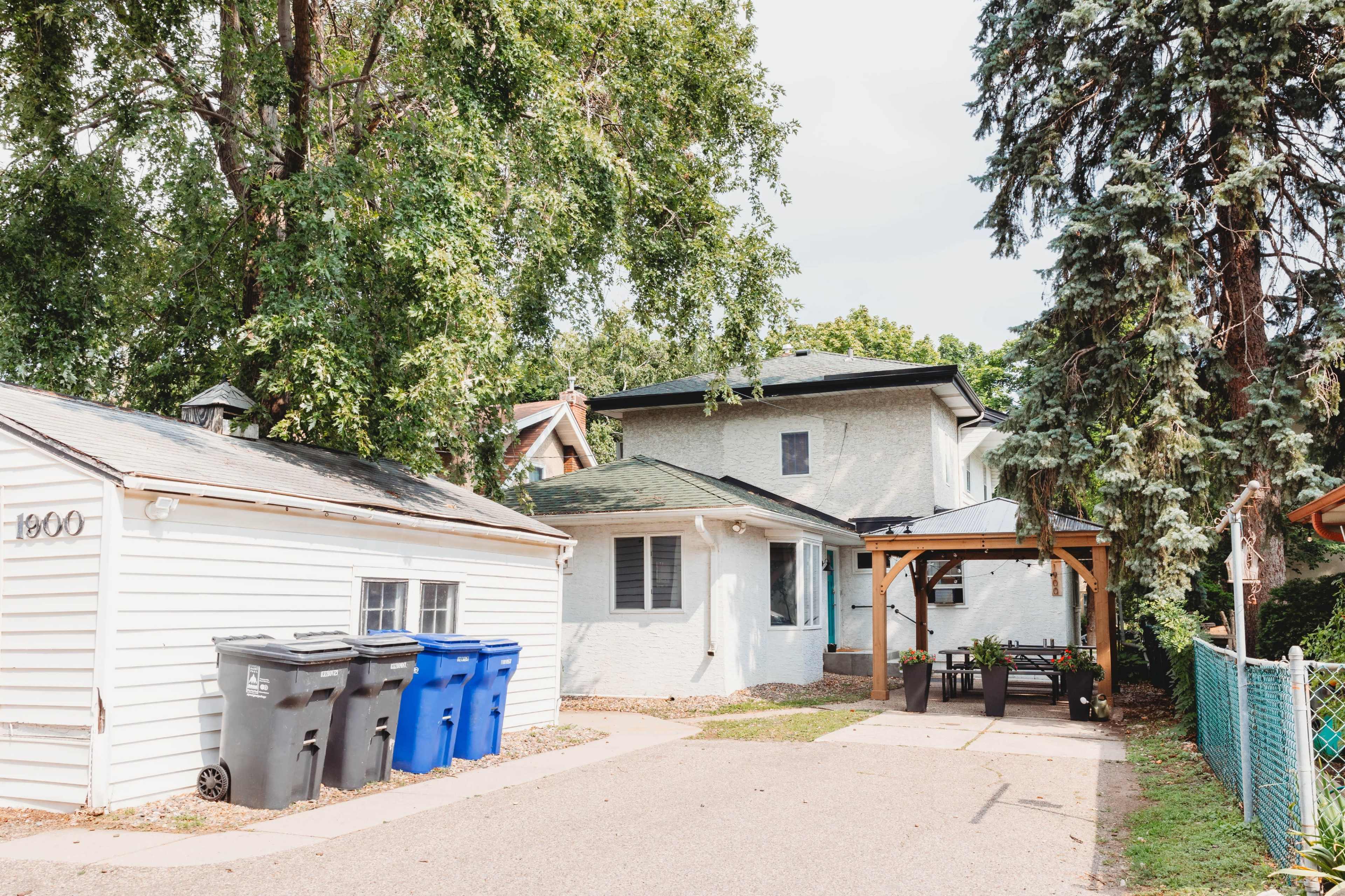 The image shows a residential property with a white garage, multiple blue trash bins, and a small patio area with a pergola.