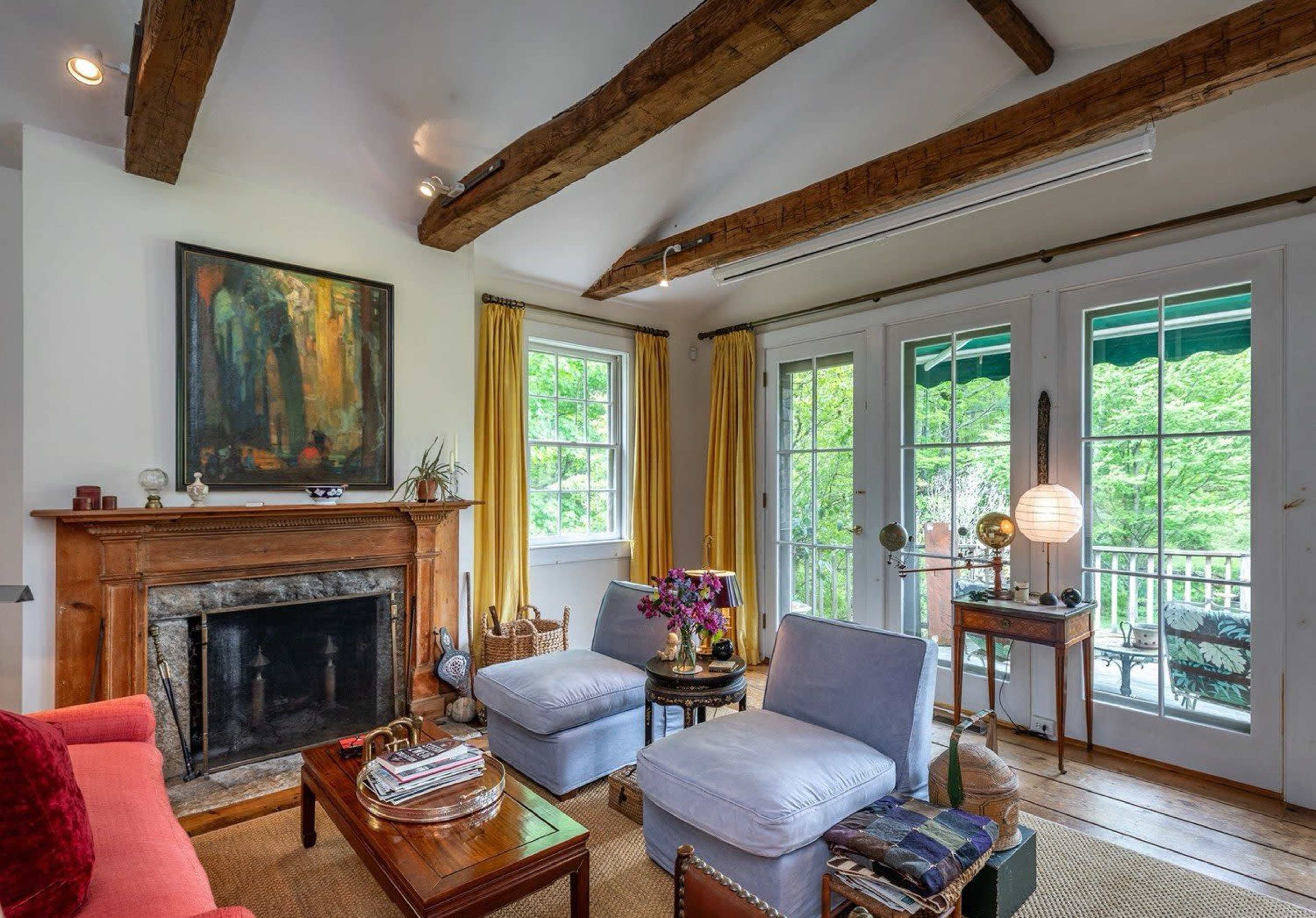 The image shows a cozy living room featuring a fireplace, two blue chairs, a wooden coffee table, and large windows with yellow curtains that open to a green outdoor space.