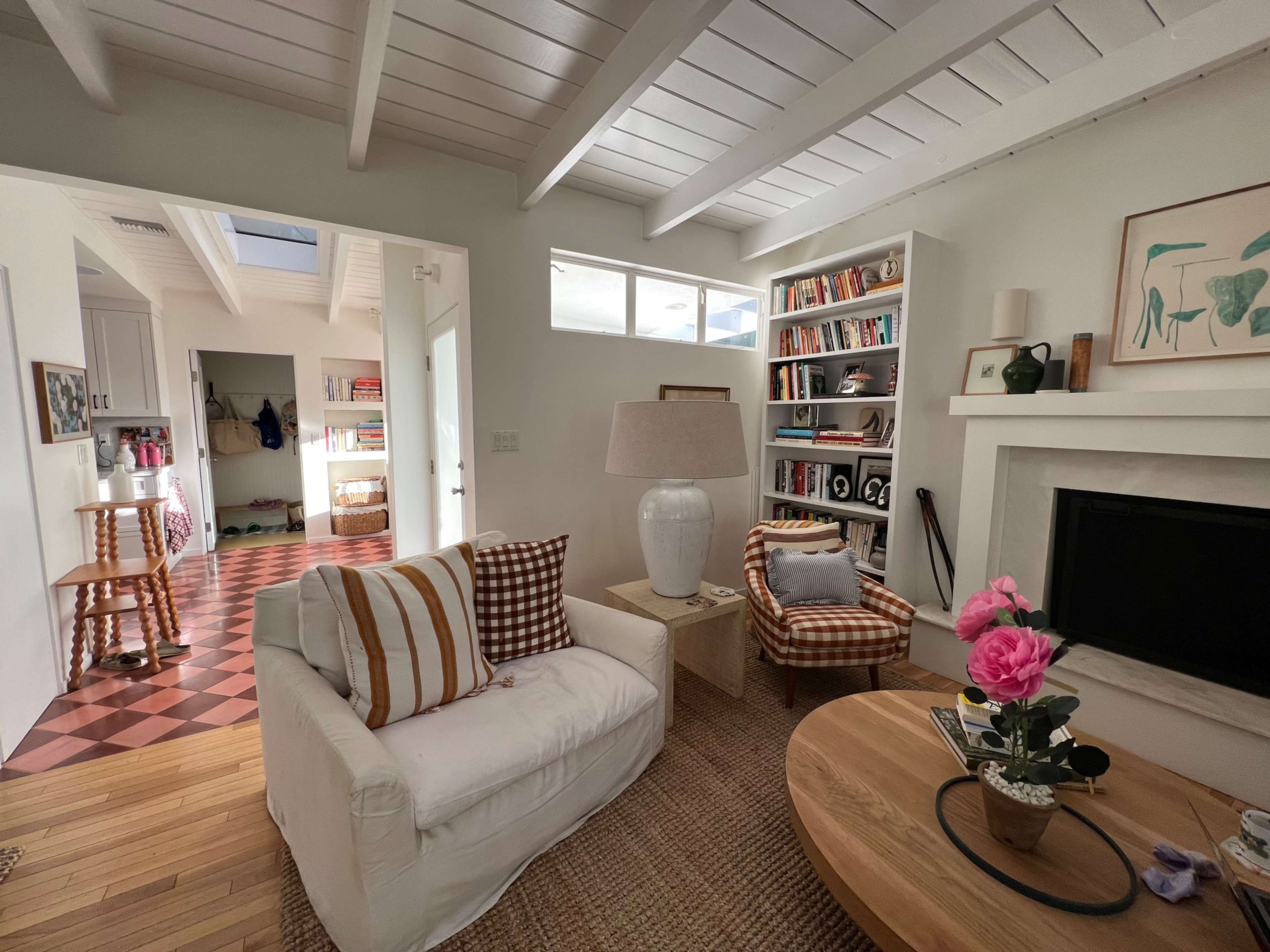 A bright living room featuring a white sofa, a patterned chair, a coffee table with flowers, and a bookshelf filled with books.