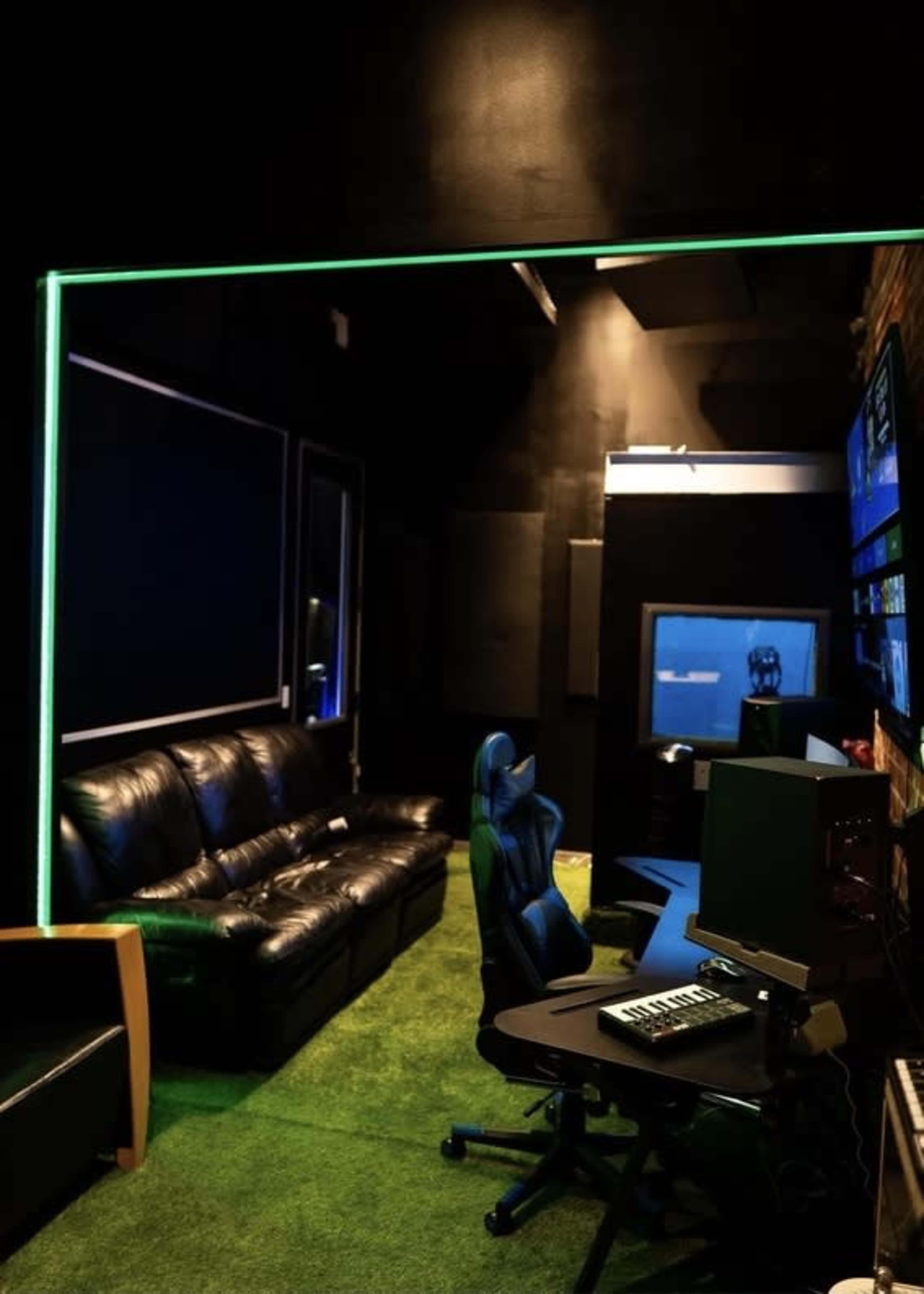 The image shows a dark room with black walls, a green LED border, a leather sofa, office chairs, and computer stations.