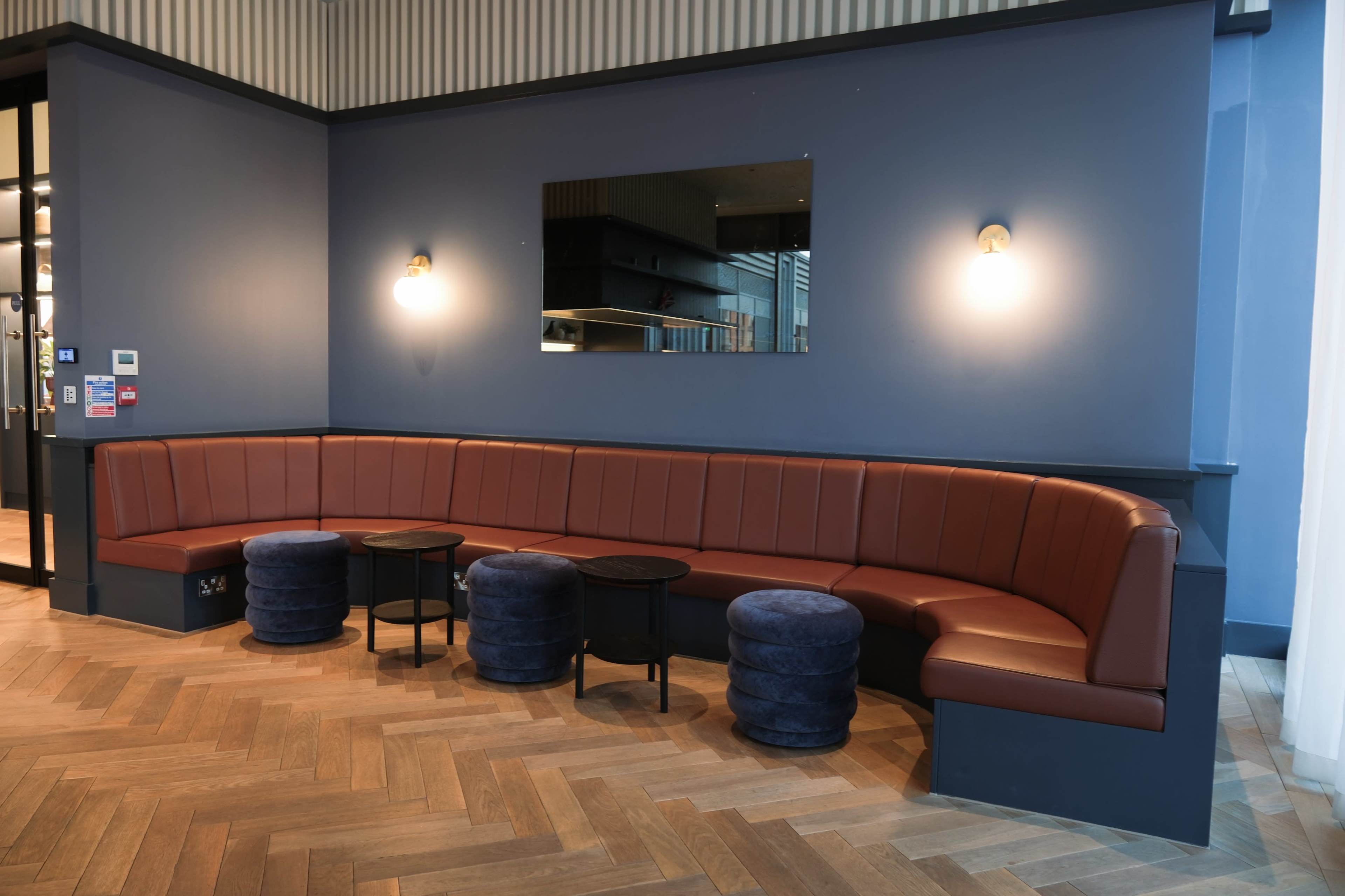 A modern seating area featuring a curved brown leather banquette against a blue wall, accompanied by small black tables and round blue stools.