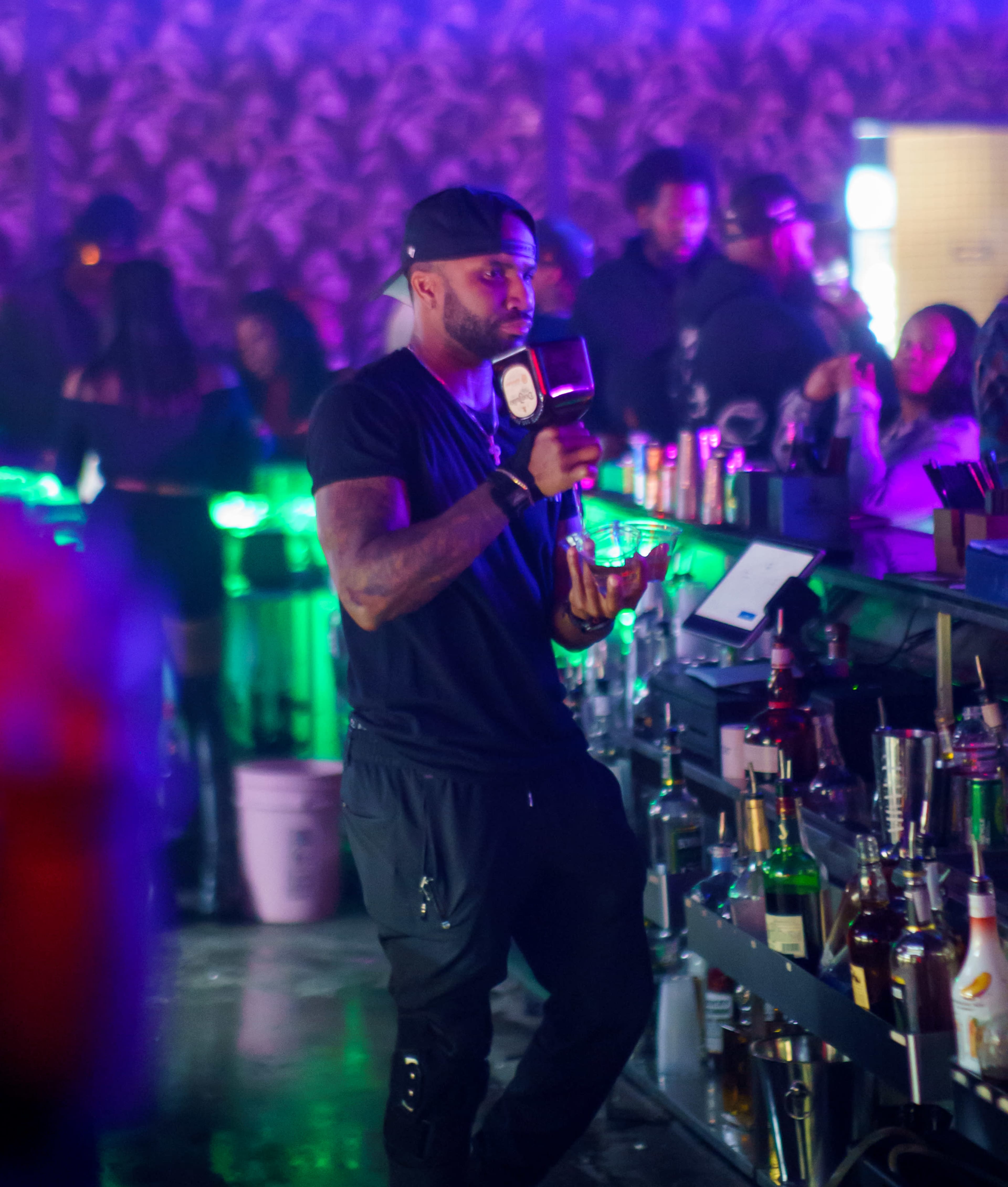 A bartender stands at the bar holding a drink in a lively nightclub setting with colorful lighting and patrons in the background.